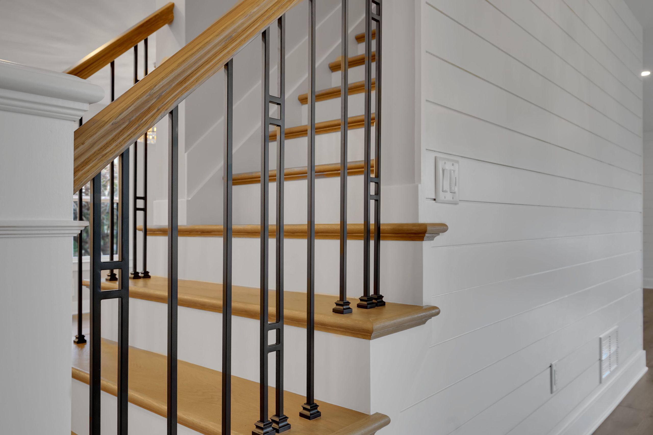 Close-up view of a wooden staircase with black metal balusters and a light wood handrail, next to a white shiplap wall in a modern, well-lit home interior.