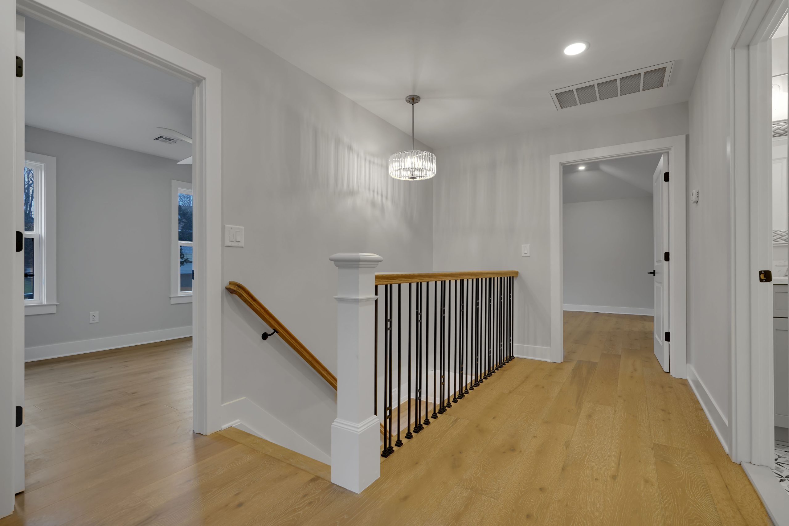 A modern hallway with light wood floors, white walls, a stairway with a black railing, and doors leading to other rooms. A round ceiling light hangs above the stairs.