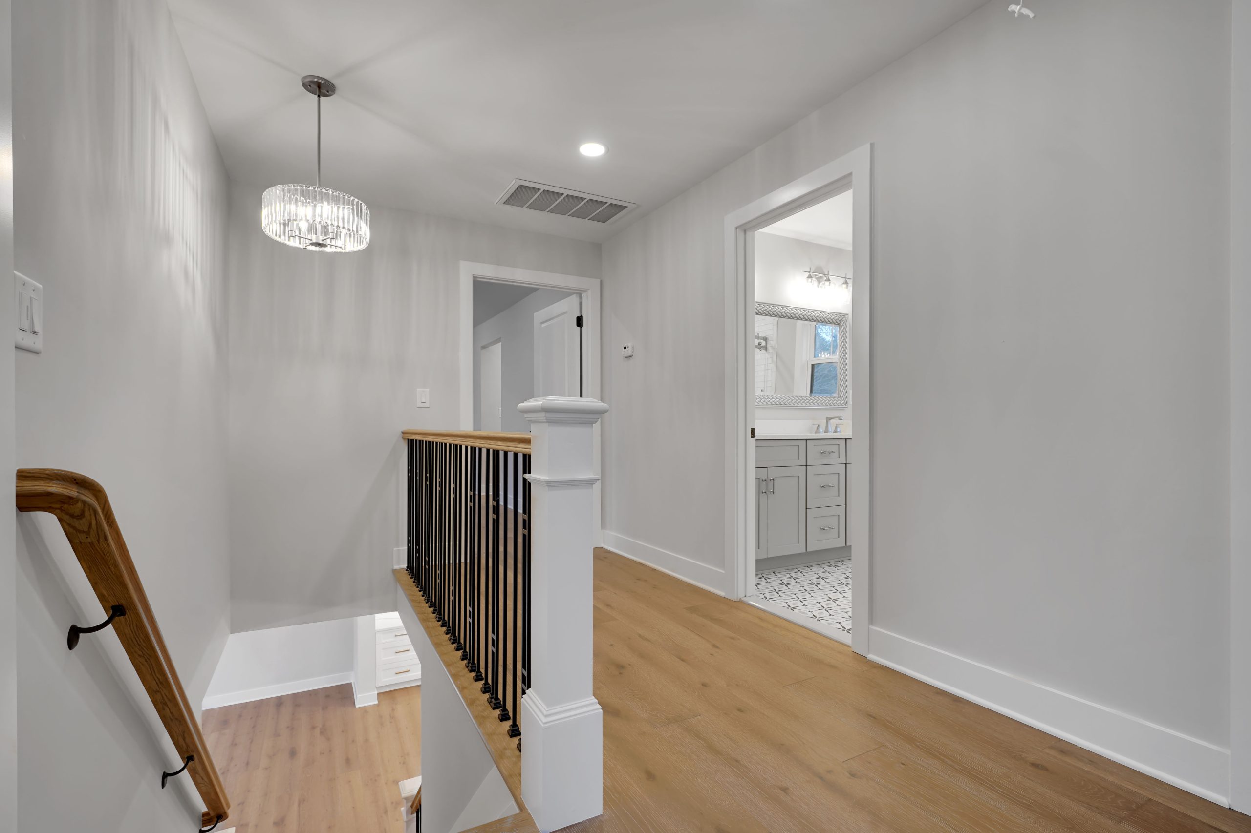 A modern, well-lit hallway with light gray walls, wooden floors, a black and wood stair railing, and a crystal light fixture; a bathroom with white cabinetry is visible through an open door.