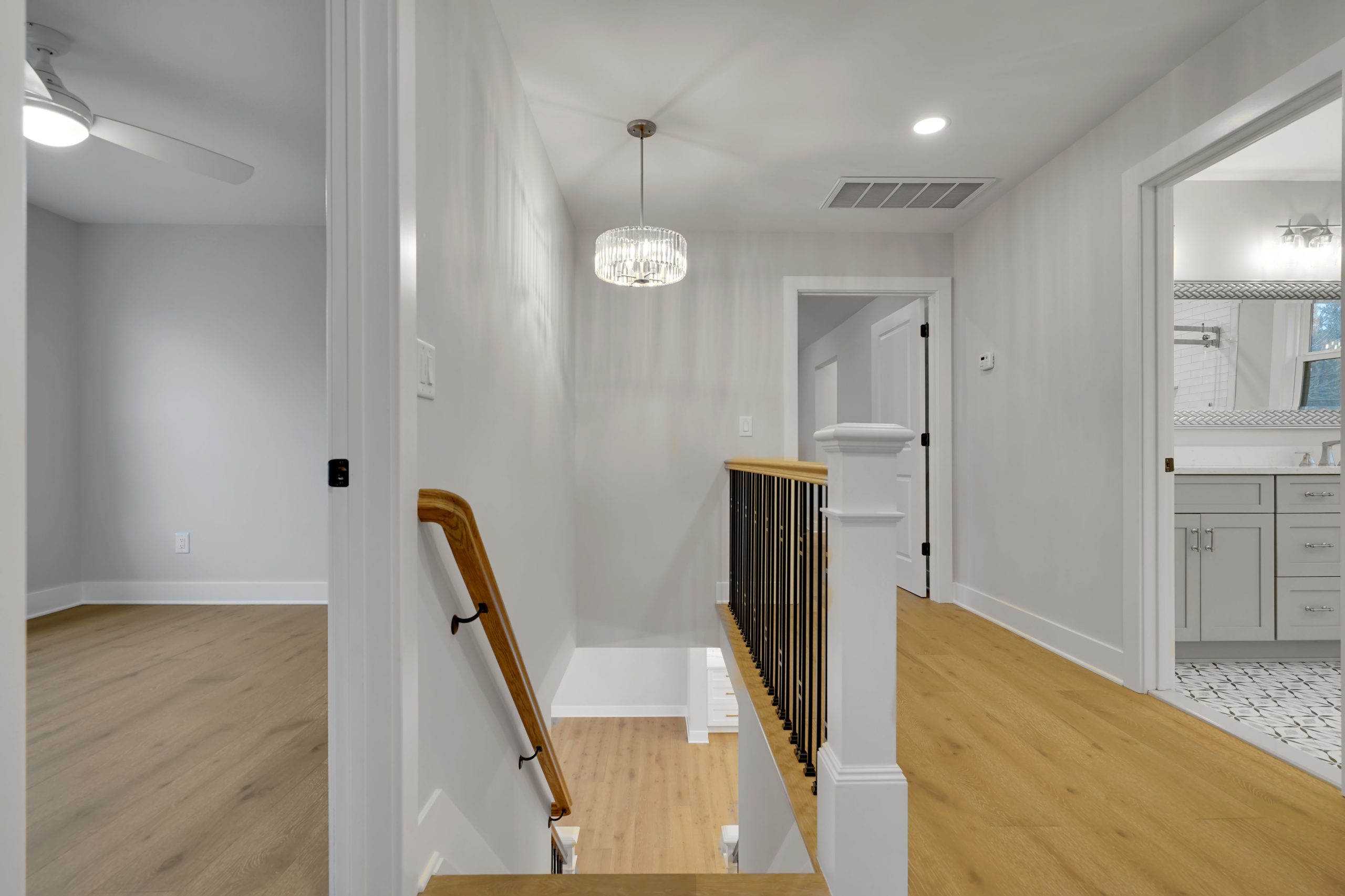 A hallway with light wood floors, white walls, a modern chandelier, and a black handrail on the staircase. To the right is a bathroom with a white vanity, and to the left, a room with a ceiling fan.