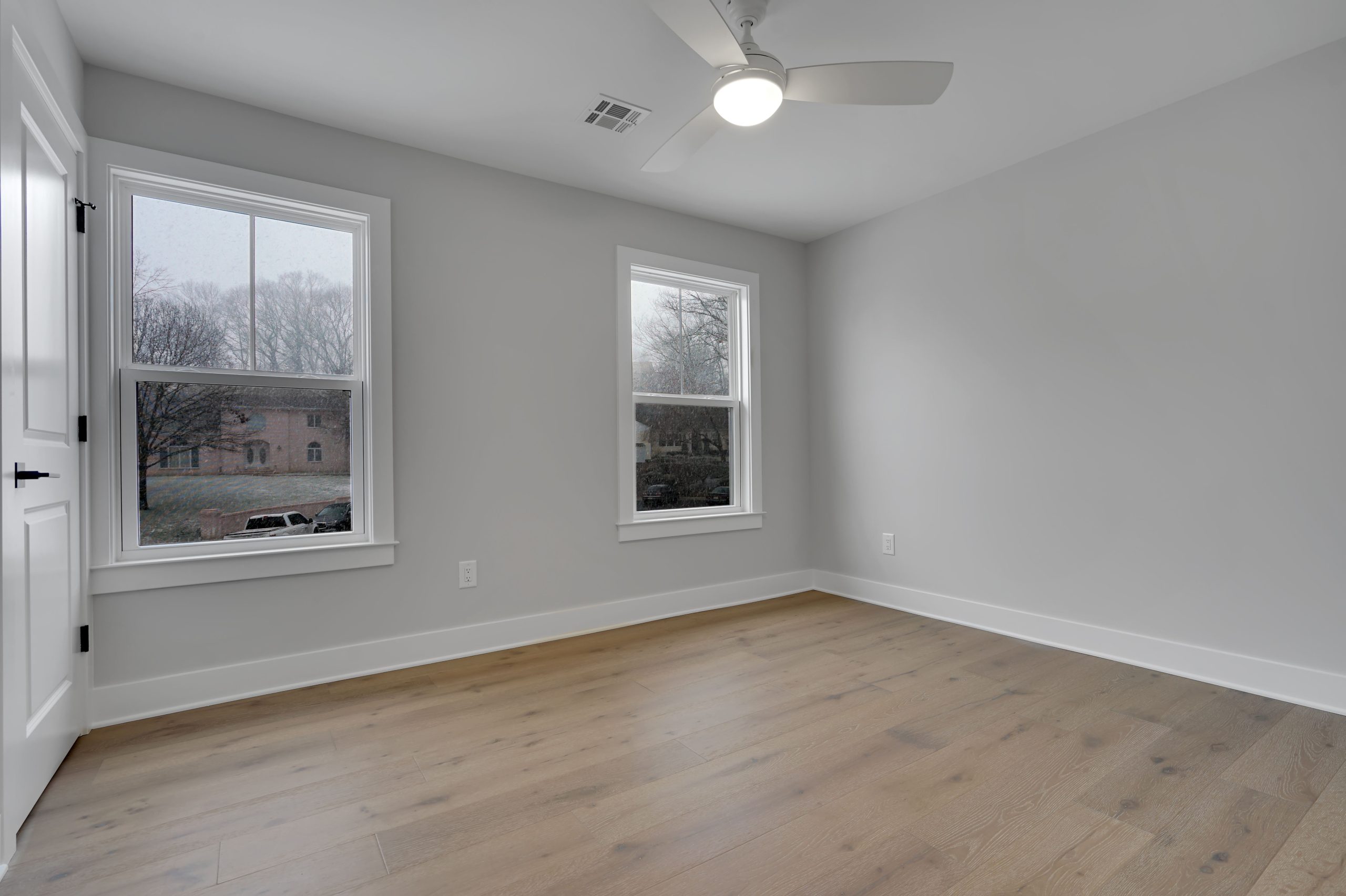 A bright, empty room with light wood flooring, light gray walls, two large windows, a ceiling fan with a light, and white trim. Trees and a building are visible outside the windows.