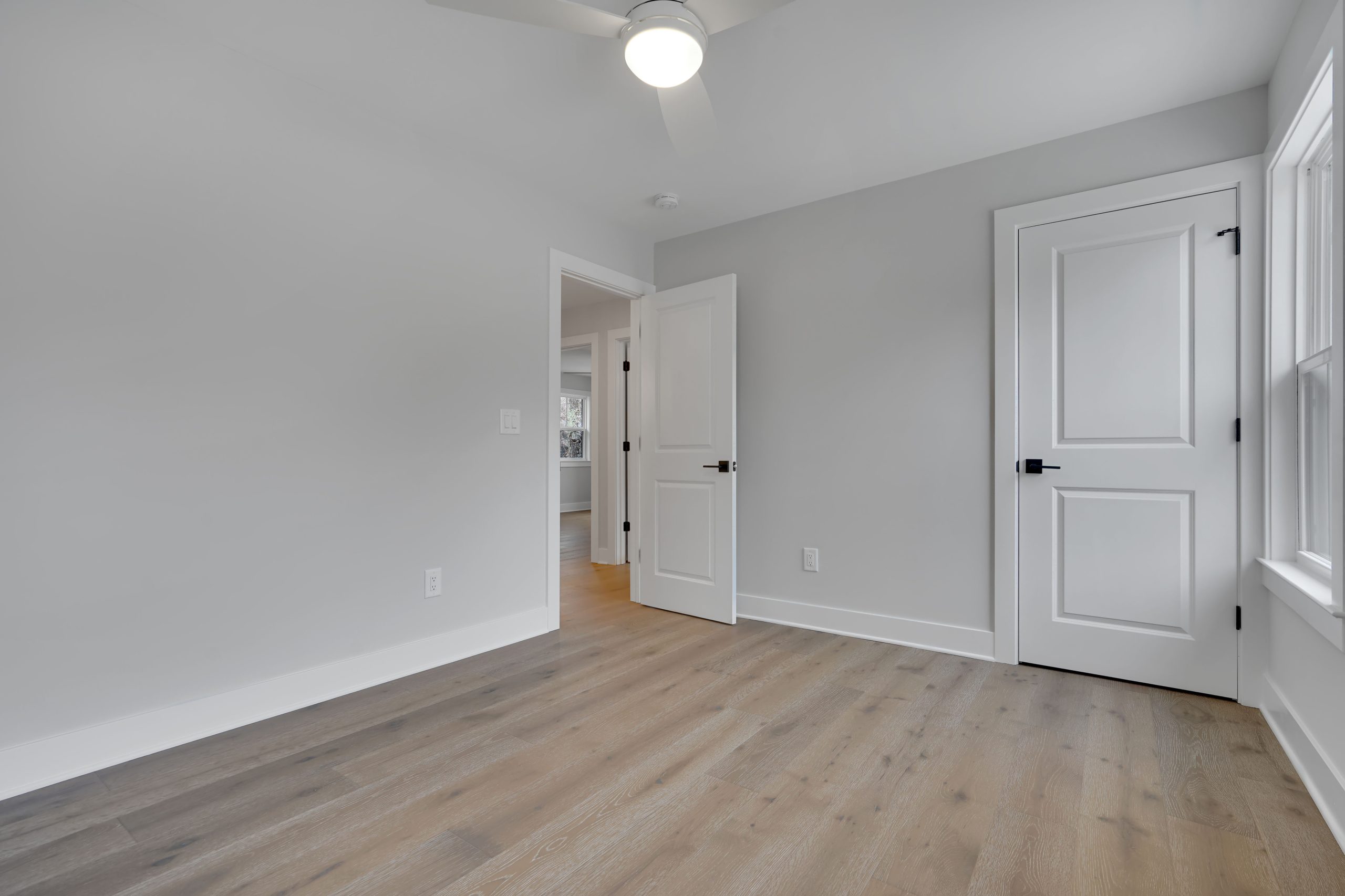 A bright, empty room with light gray walls, wood flooring, a ceiling fan with light, two white doors, and a large window allowing natural light to enter.