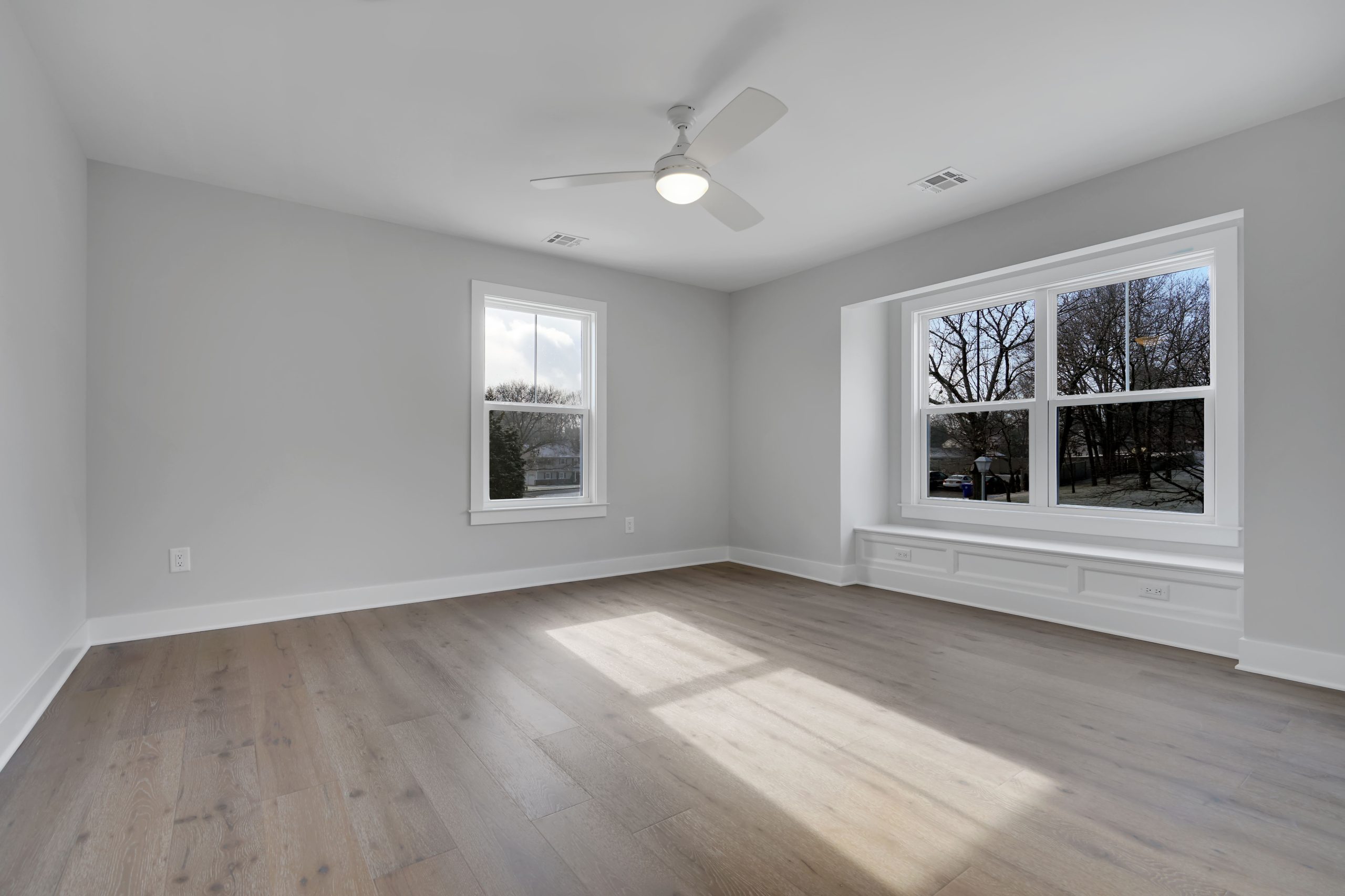 A bright, empty room with light wood floors, light gray walls, a ceiling fan, and two large windows letting in natural light. One window has a wide ledge, and outside trees are visible.