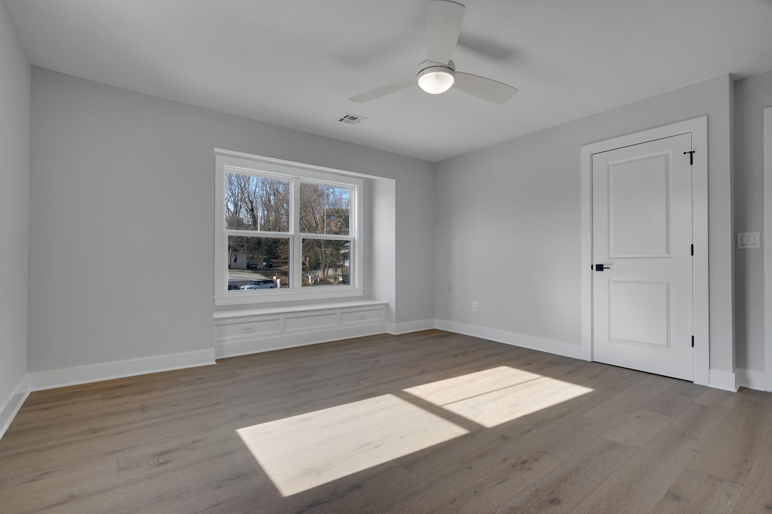 Bright, empty room with light-gray walls, large window with outdoor view, wood flooring, white door, ceiling fan, and built-in window seat beneath the window. Sunlight streams through the window, casting shadows on the floor.