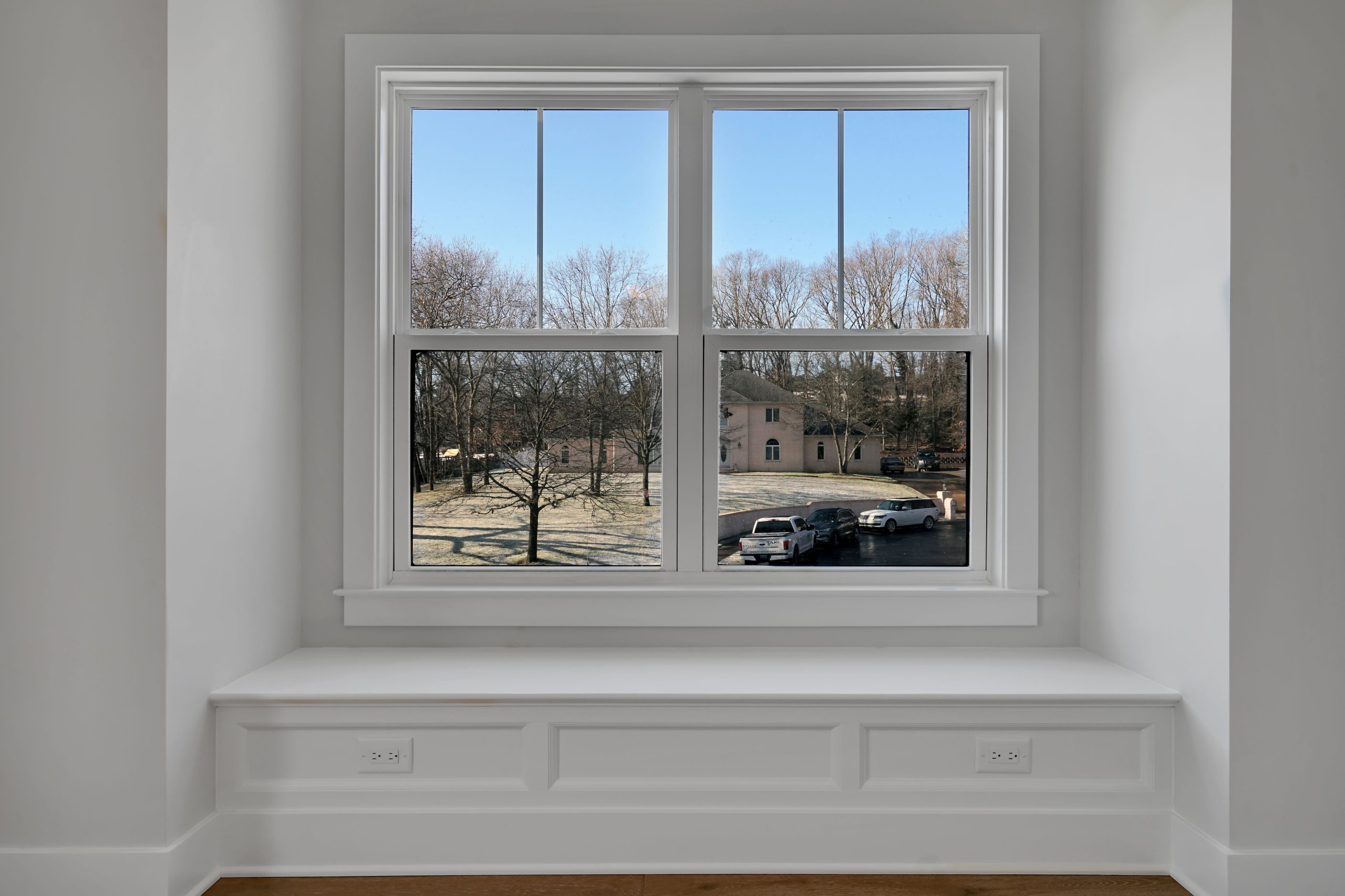 A white window seat with built-in outlets sits beneath a large window, offering a view of a suburban neighborhood with leafless trees, a house, and parked cars under a clear blue sky.