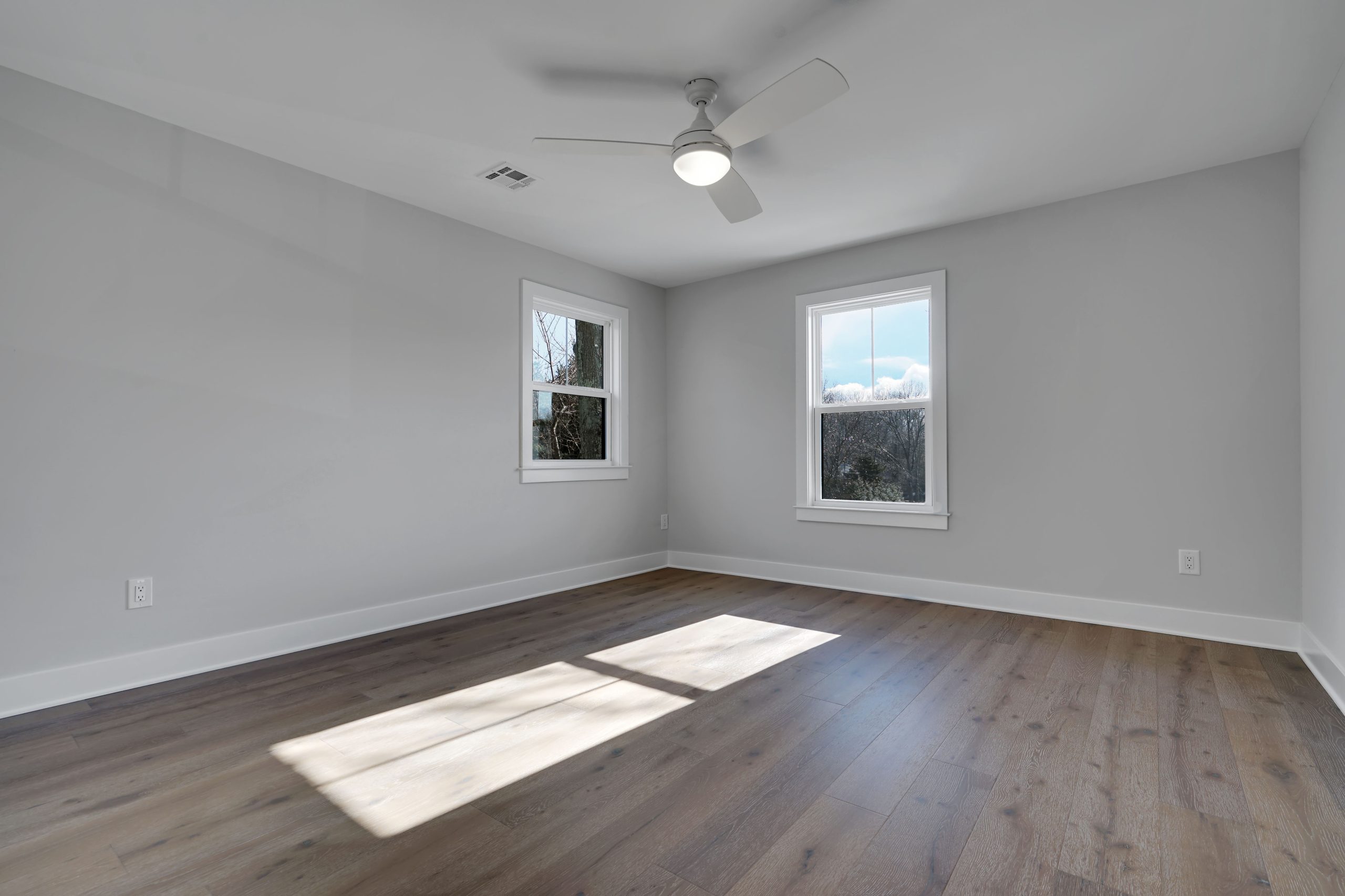 A bright, empty room with light gray walls, two windows, wood flooring, a white ceiling fan with a light, and sunlight streaming in through the windows onto the floor.