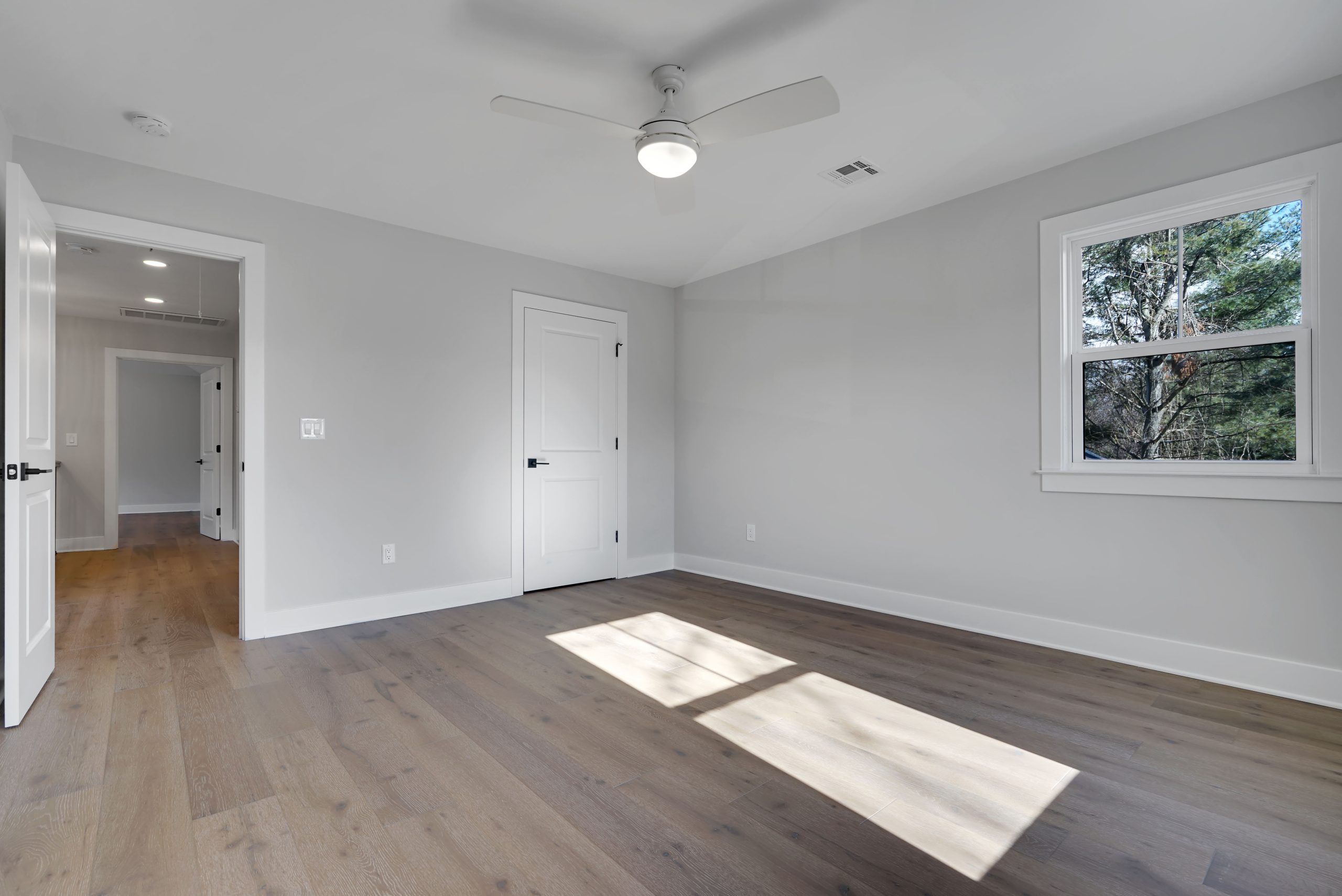 Bright, empty bedroom with light wood floors, white walls, a ceiling fan, two closed white doors, and a window with natural light streaming in and greenery visible outside.