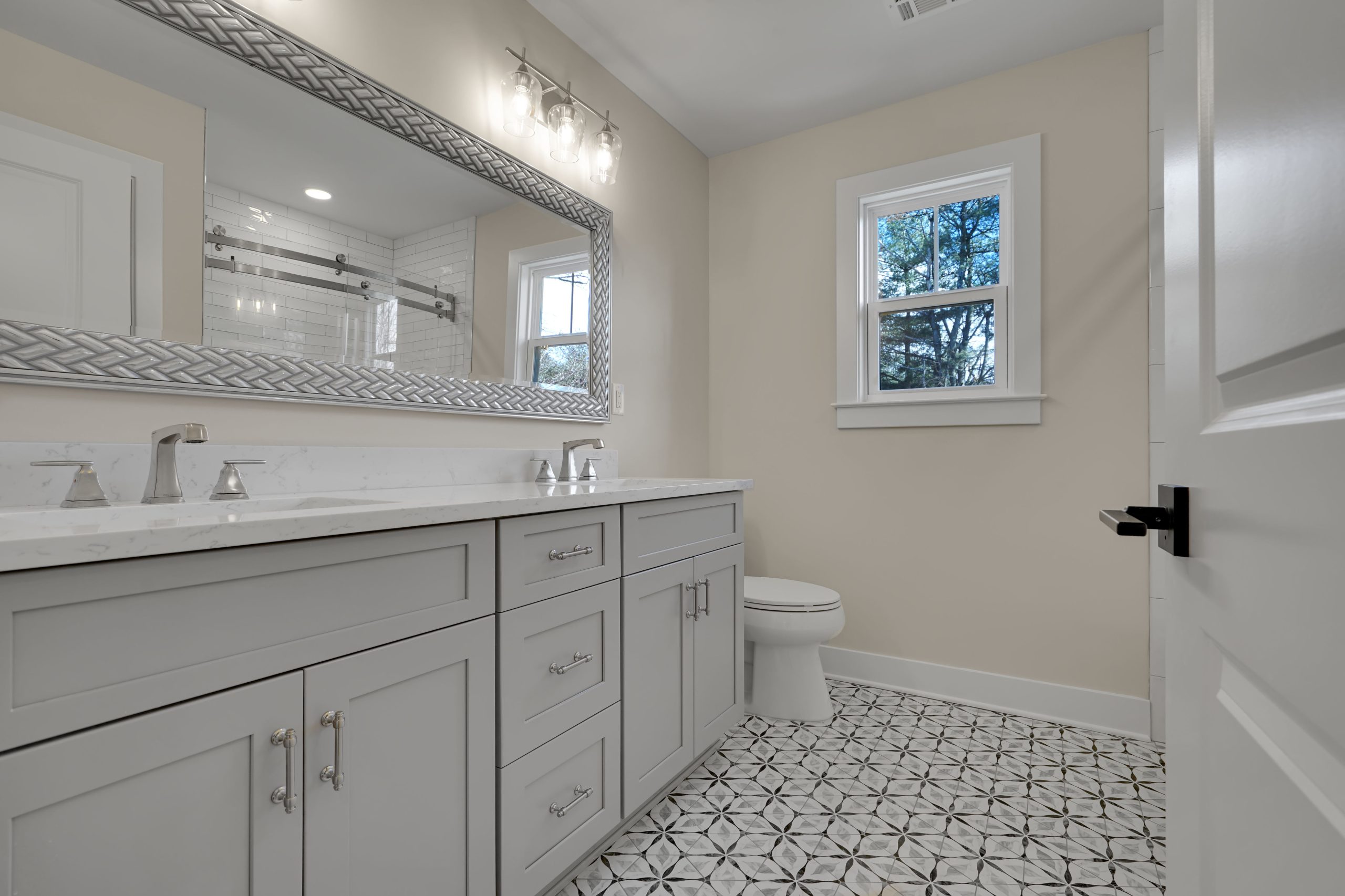 Modern bathroom with a large double-sink gray vanity, marble countertop, patterned tile floor, wall mirror, toilet, window, and soft beige walls under bright overhead lighting.