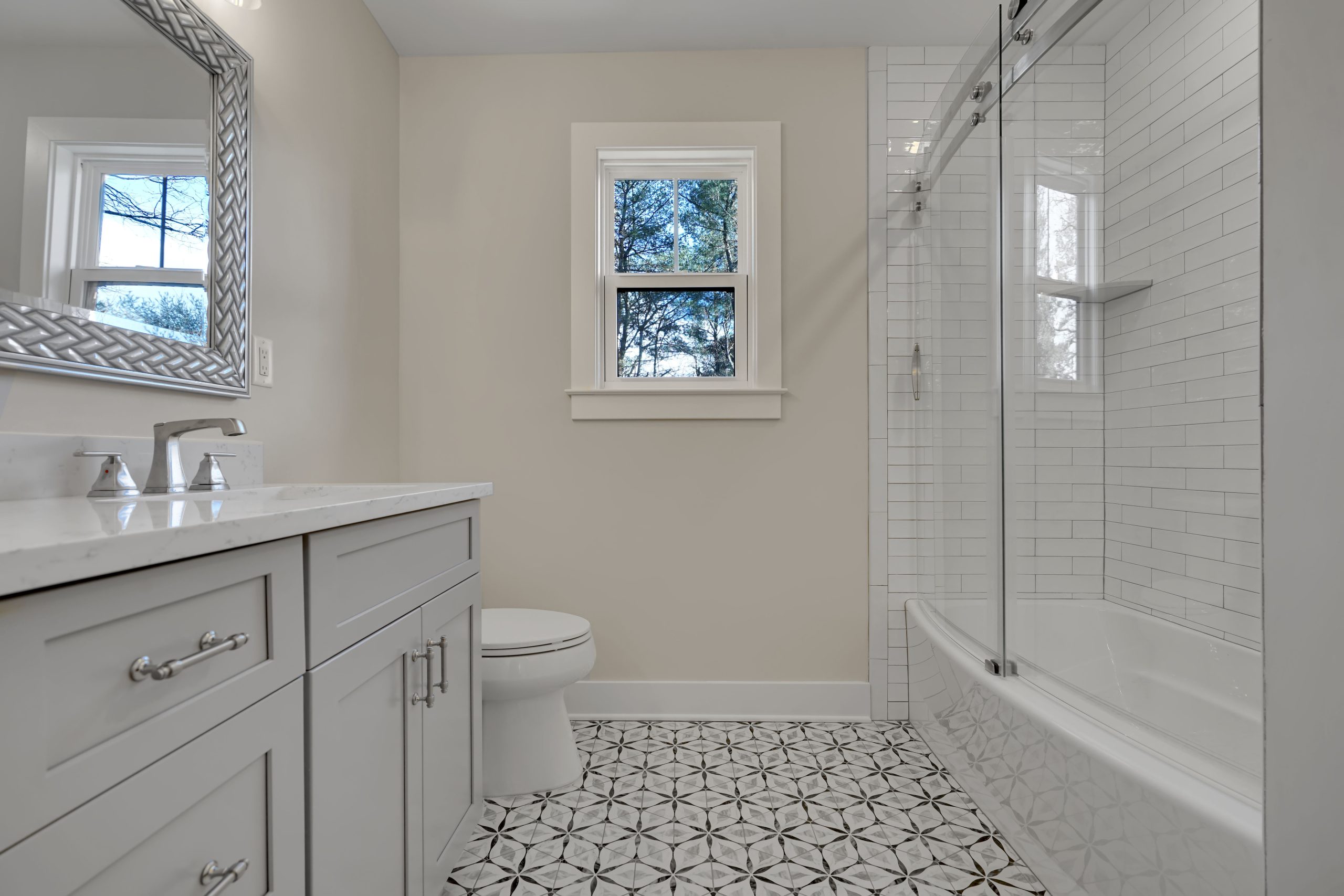 Modern bathroom with white cabinets, a large mirror, patterned tile floor, toilet, and a white bathtub with glass sliding doors and subway tile walls. A window lets in natural light above the toilet.