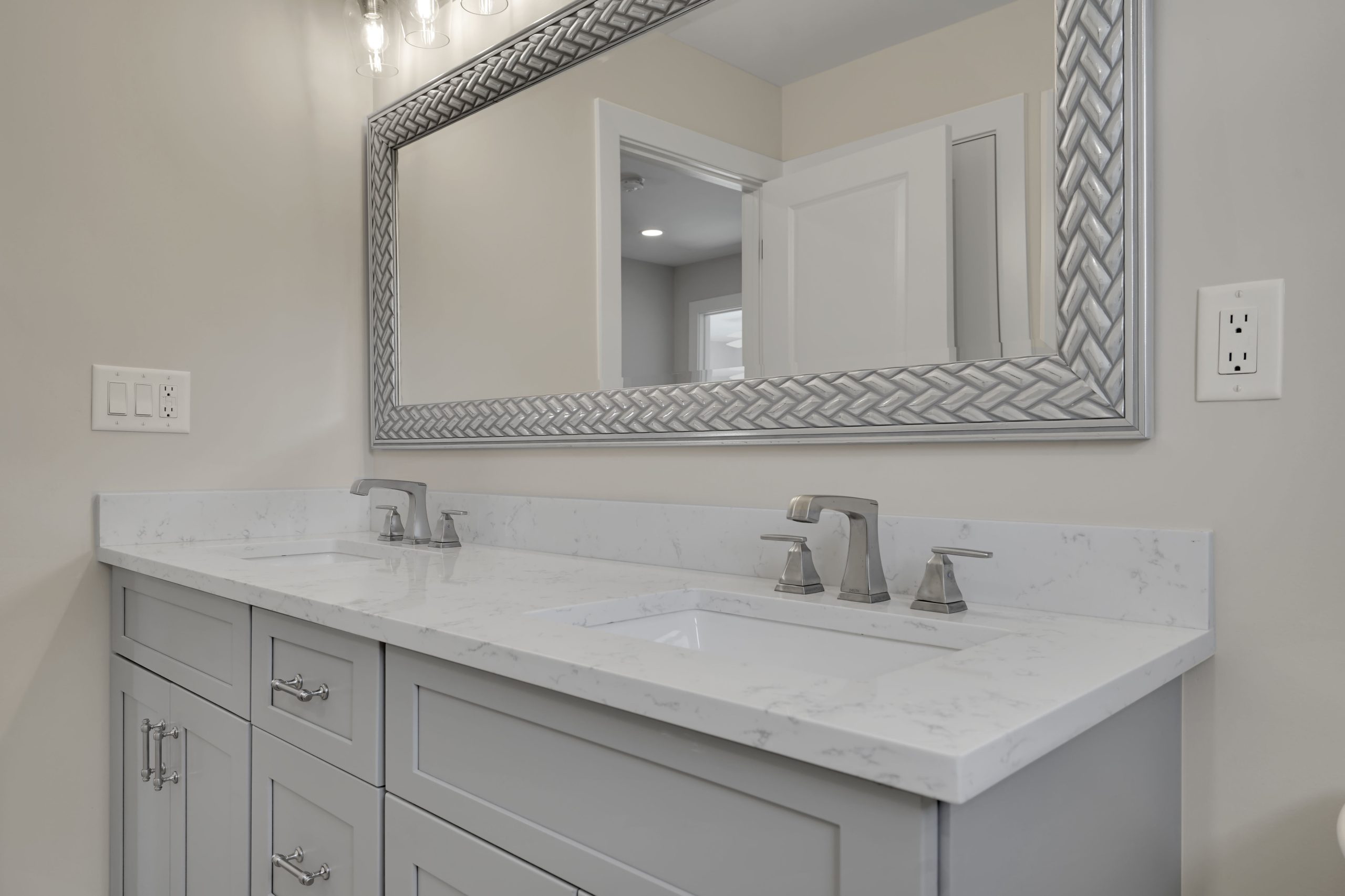 Modern bathroom with a double vanity, gray cabinets, marble countertop, two sinks with silver faucets, and a large decorative mirror above. Neutral walls and white trim complete the clean look.