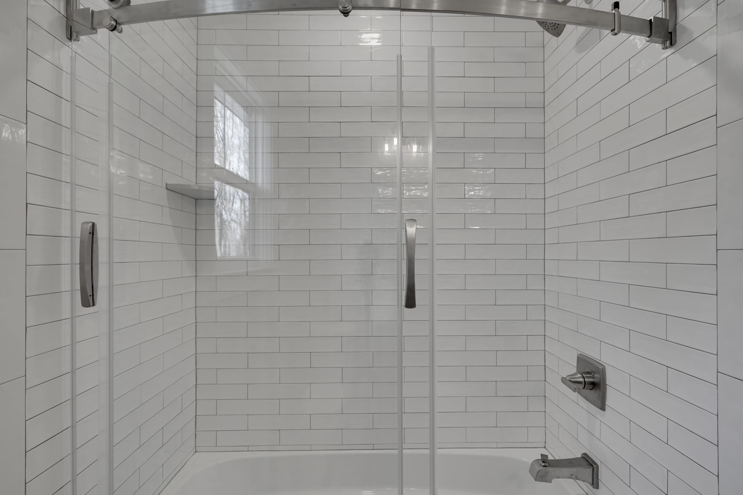 A modern bathtub and shower with white subway tile walls, a curved glass sliding door, chrome fixtures, and a small corner shelf. Natural light enters through a window reflected in the tiles.