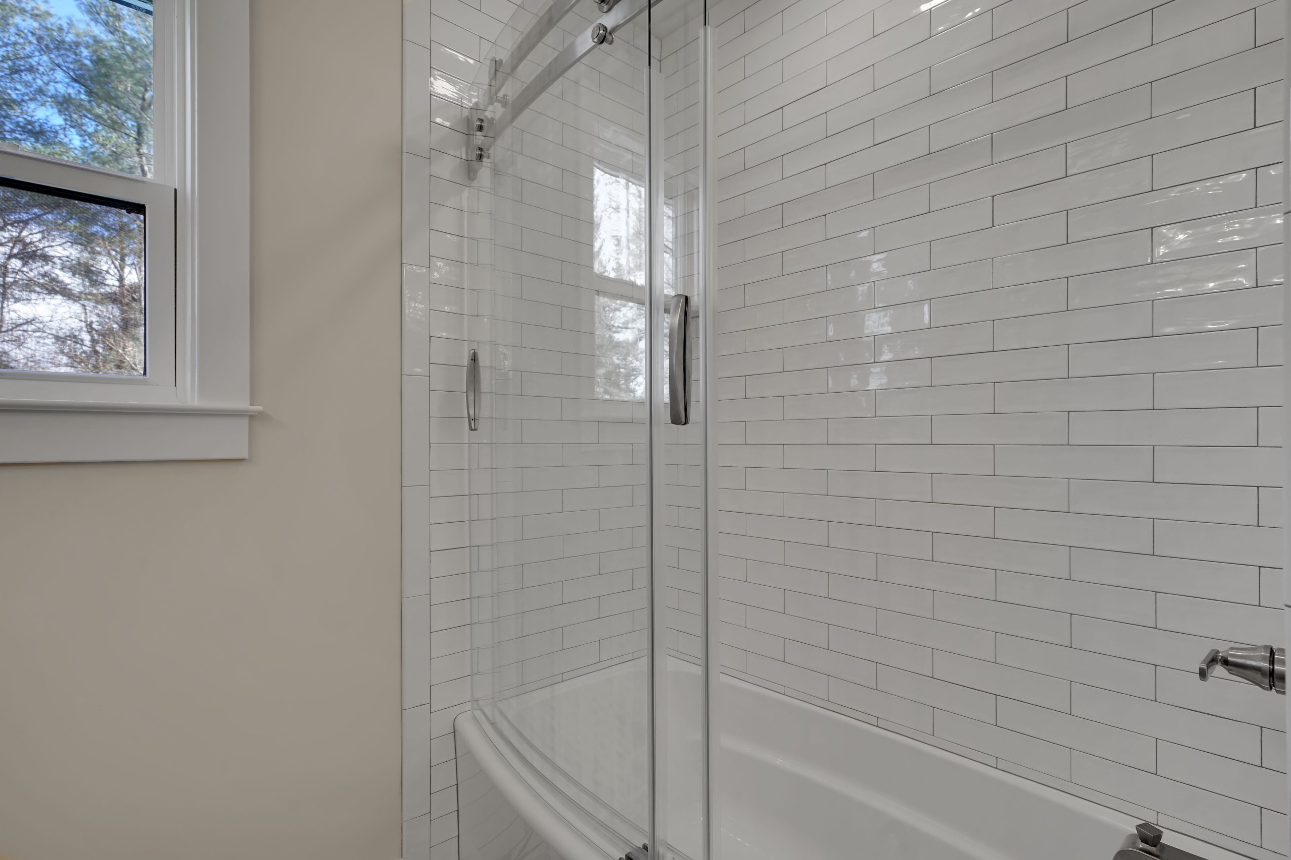 A modern bathroom with white subway tile walls, a glass shower door, a white bathtub, and a window letting in natural light.