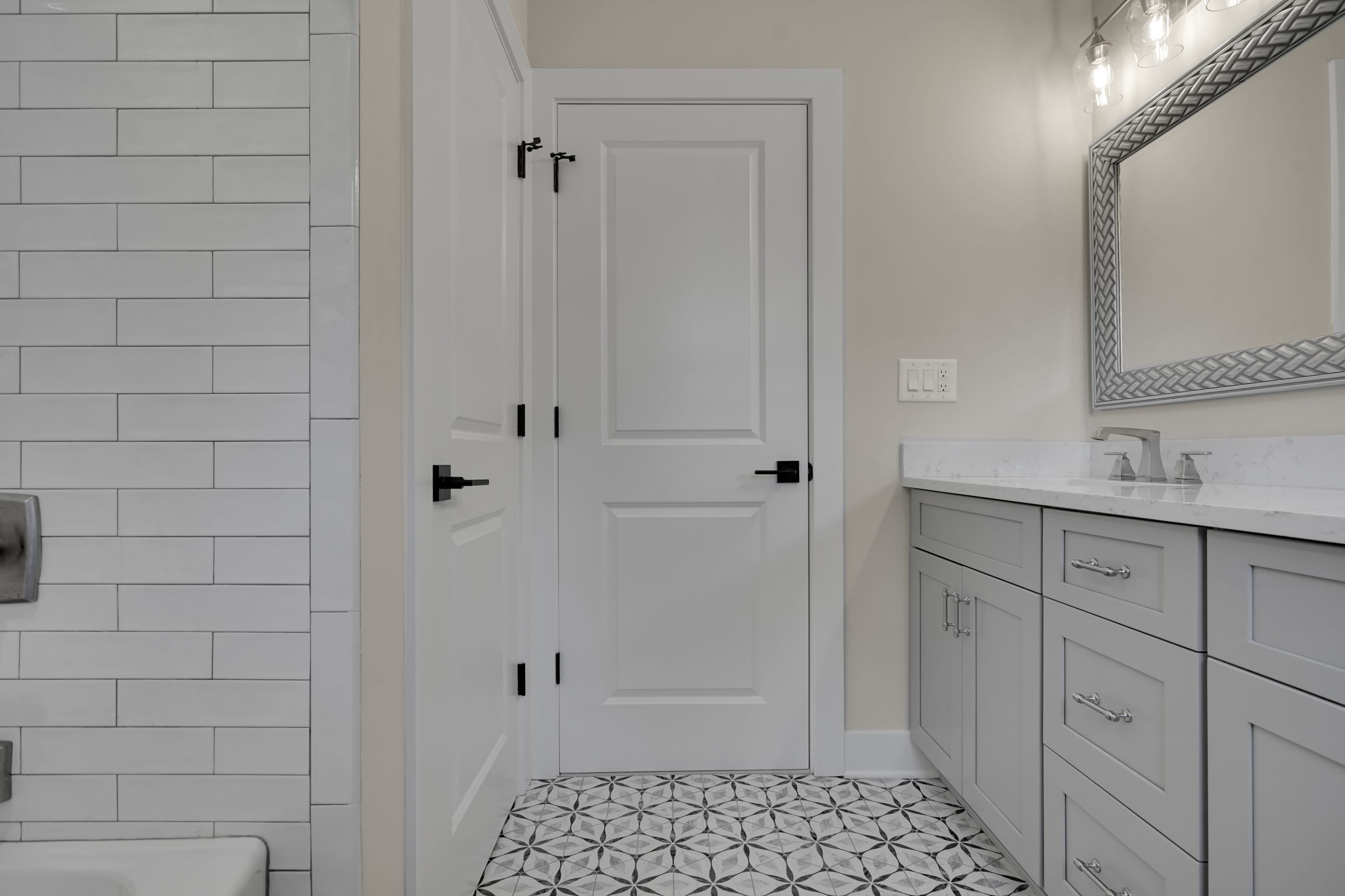Modern bathroom with geometric patterned tile floor, light gray vanity with dual sinks, white countertop, large framed mirror, and white doors. Subtle lighting fixtures and white subway tiles are visible on the left.