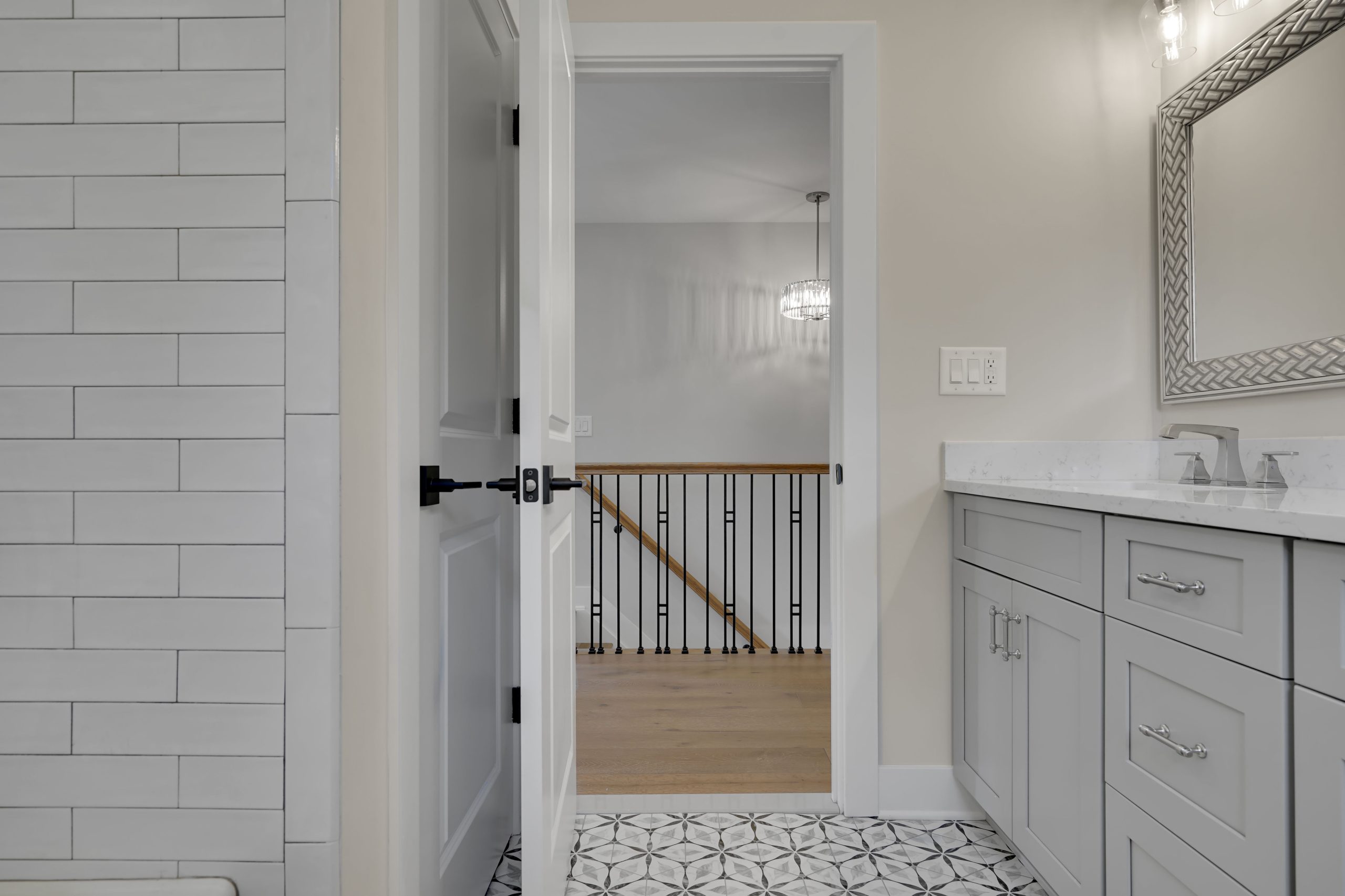 A modern bathroom with gray cabinets, white countertop, patterned tile floor, and a large mirror. The door is open, showing a hallway with wooden flooring and a black iron stair railing.