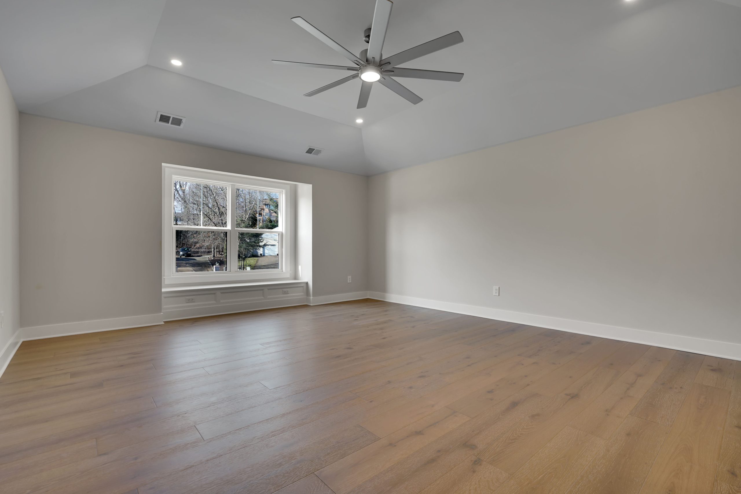 A spacious, empty room with light gray walls, wide-plank wood flooring, a large window with outdoor view, ceiling fan with lights, and recessed lighting in the ceiling.