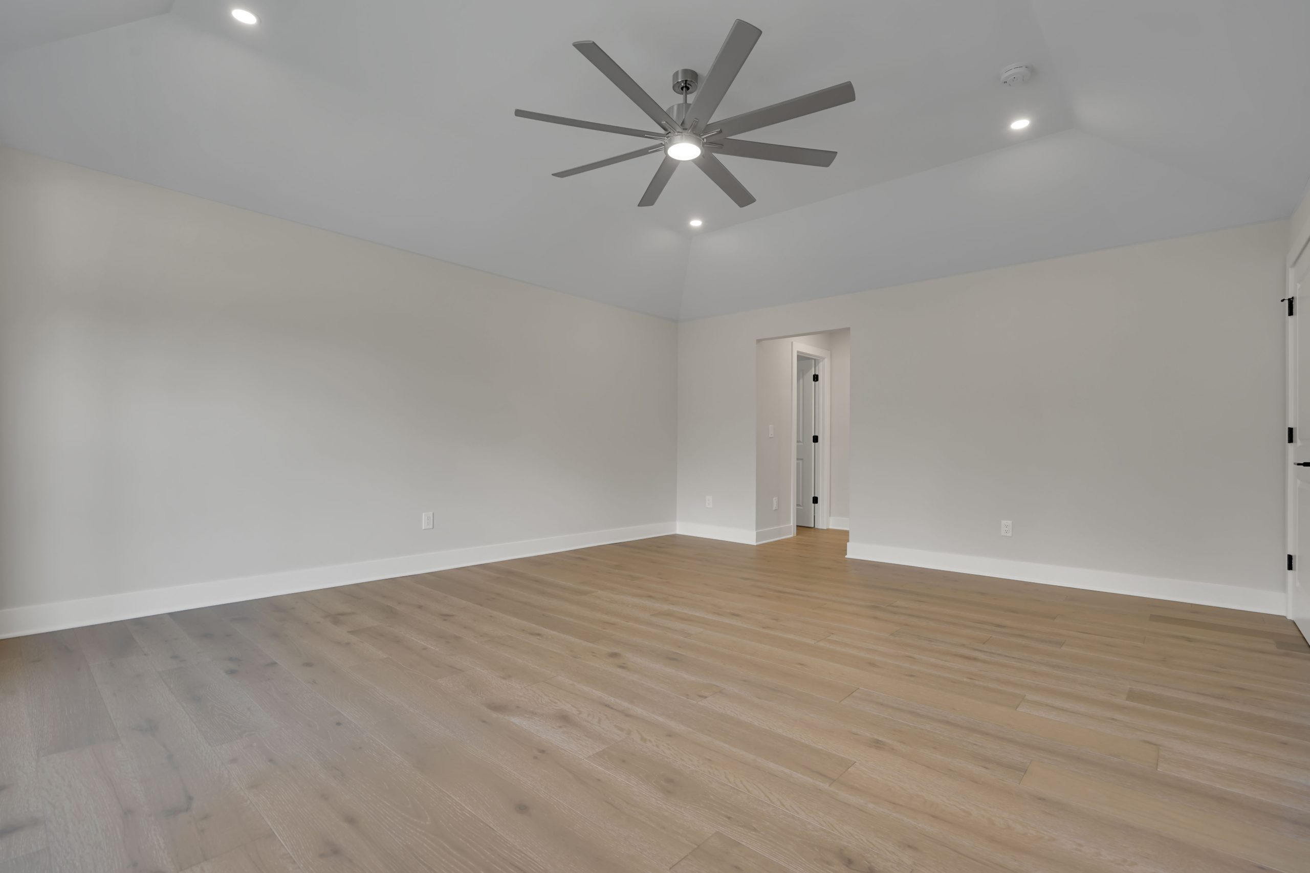 A spacious, empty room with light-colored walls, wood flooring, a large ceiling fan with multiple blades, recessed lighting, and two doorways leading to other areas.