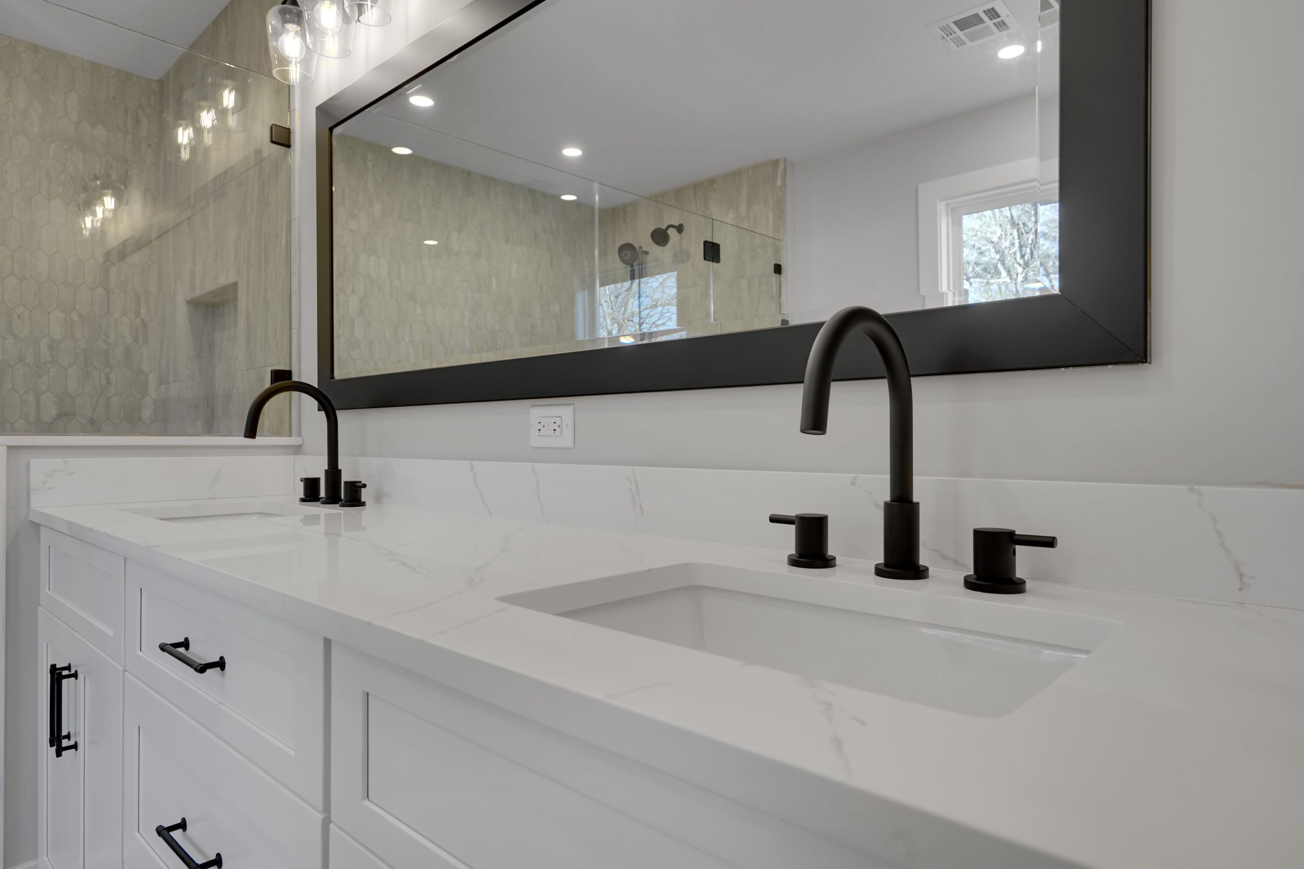 Modern bathroom with a double sink vanity, white countertop, black faucets, and a large rectangular mirror above. Light fixtures and a textured wall are reflected in the mirror.