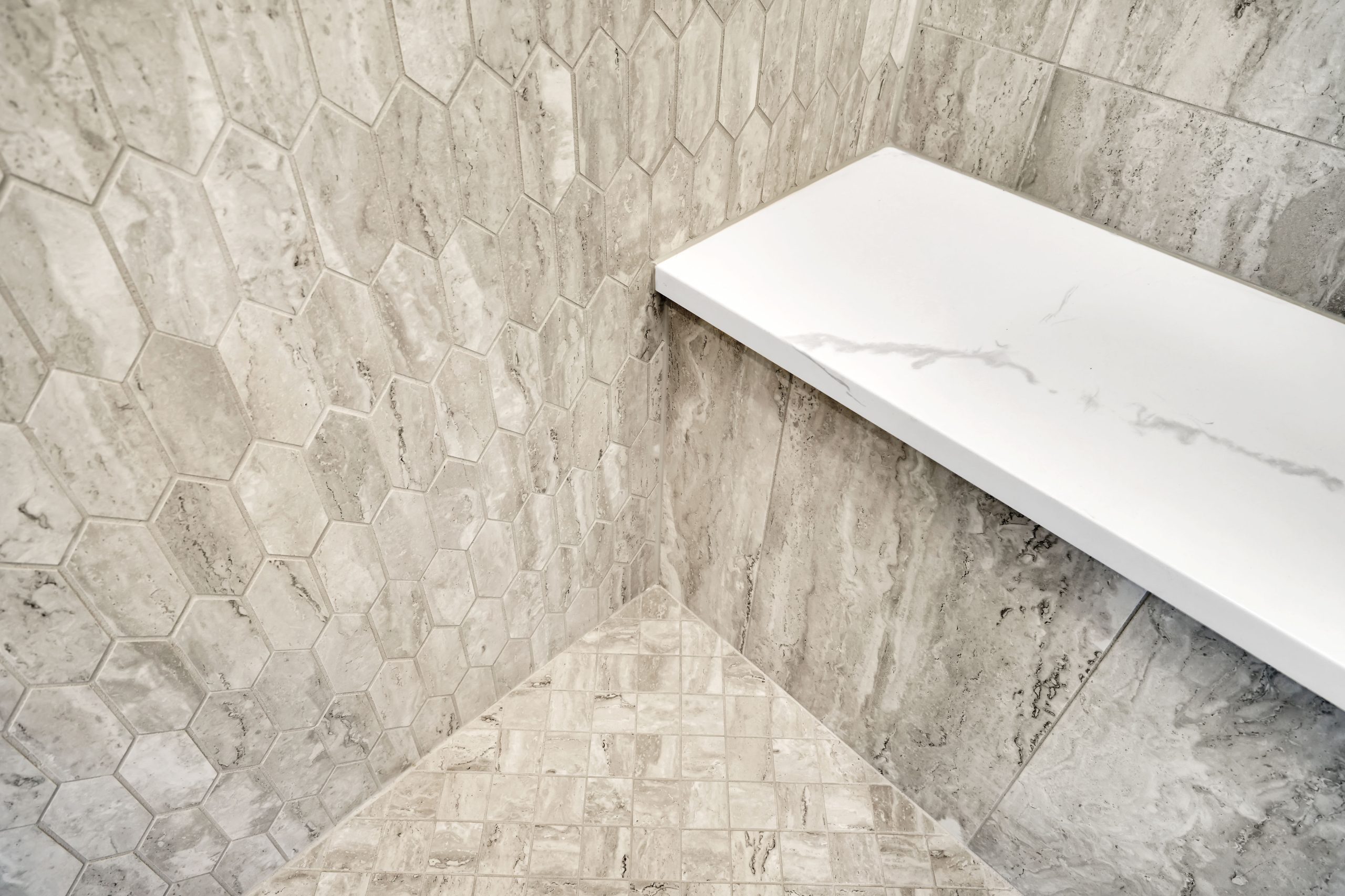 Close-up of a modern shower corner with light gray hexagon wall tiles, a white marble bench, and square stone floor tiles in neutral tones.