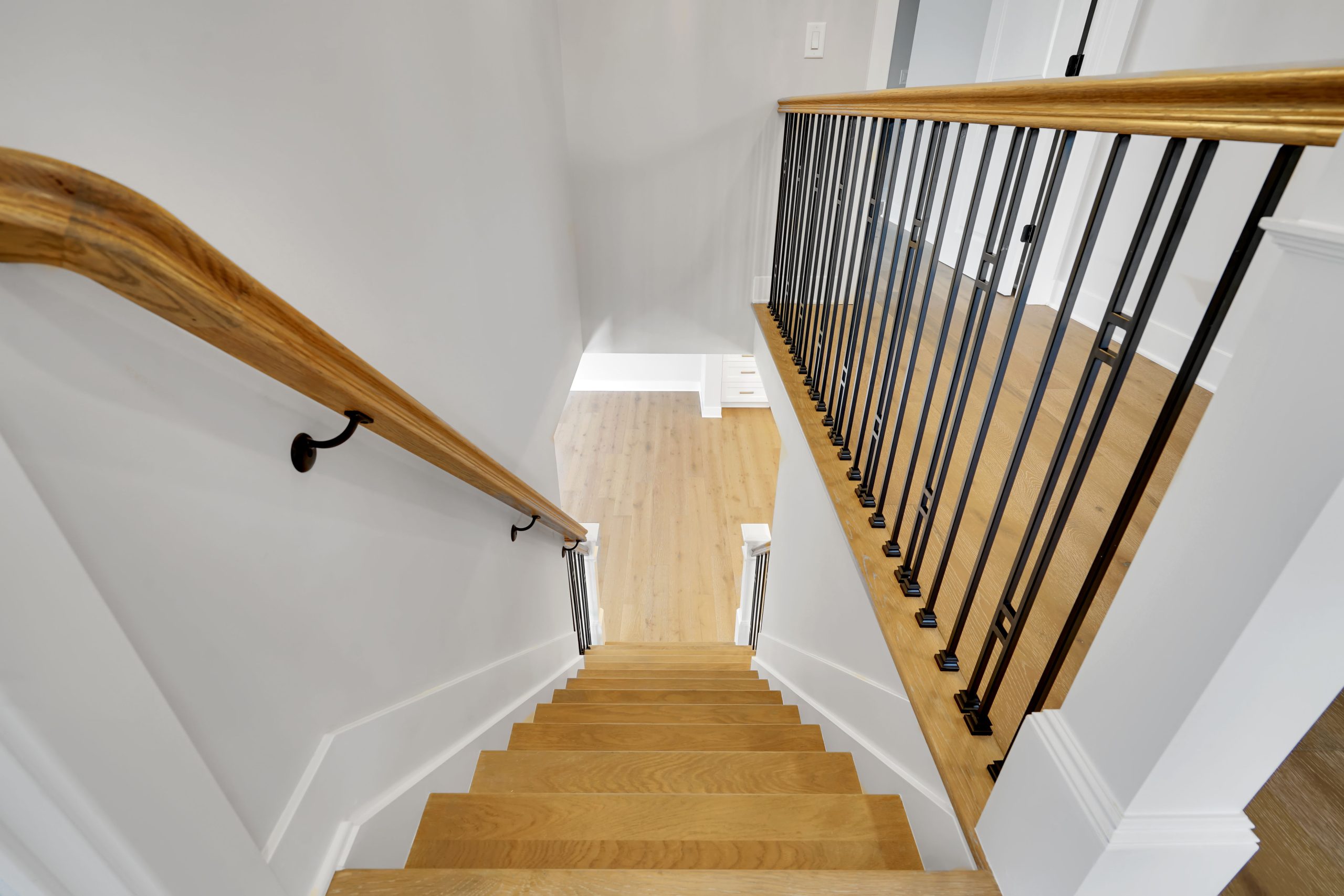 A top-down view of a wooden staircase with a black metal railing and a wooden handrail, leading down to a light-colored hardwood floor in a modern, bright interior space.