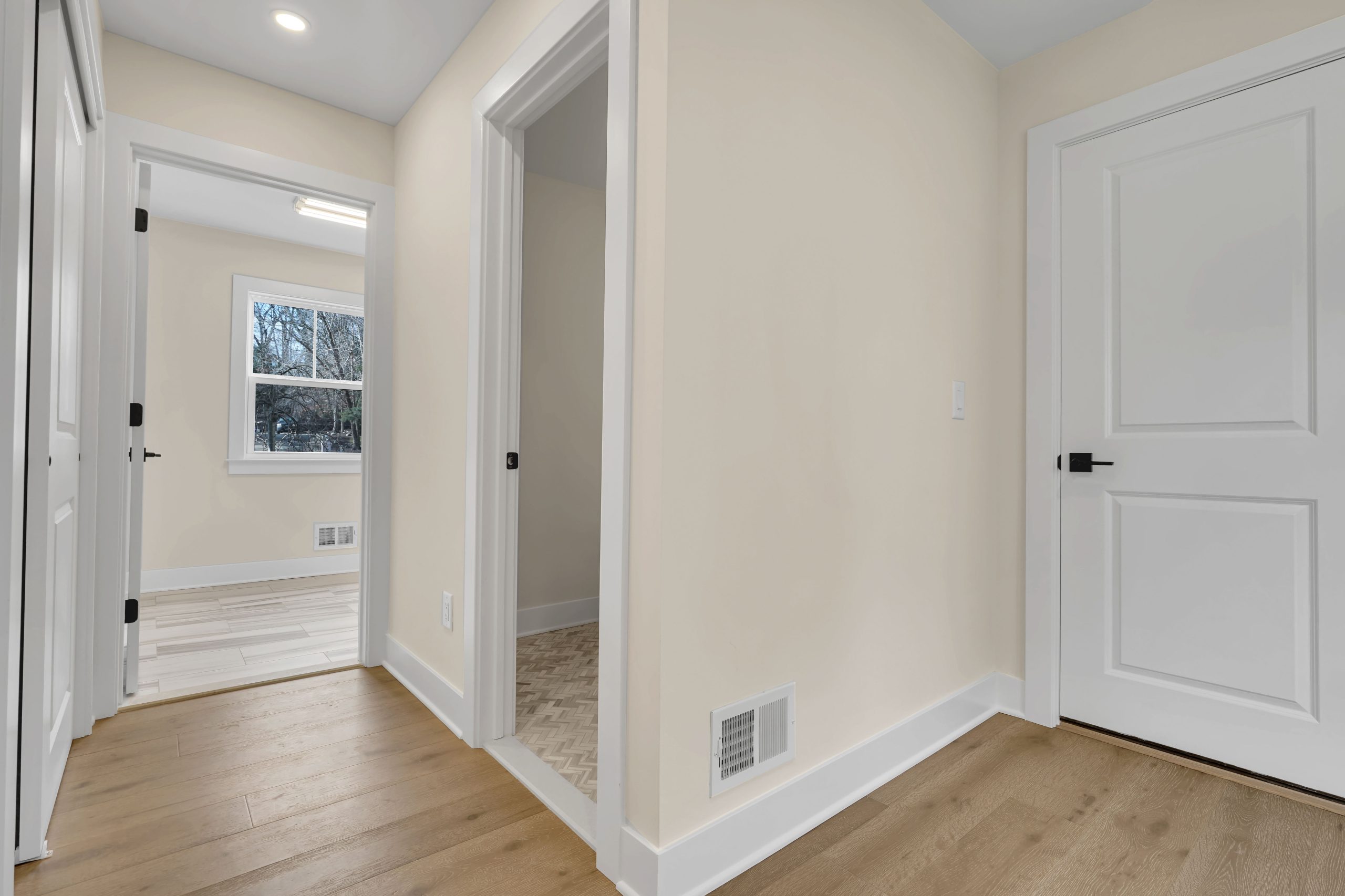 A hallway with light wood flooring, cream-colored walls, white trim, and three doorways. One doorway reveals a bright room with a window, another shows a small tiled space, and the third is a closed white door.