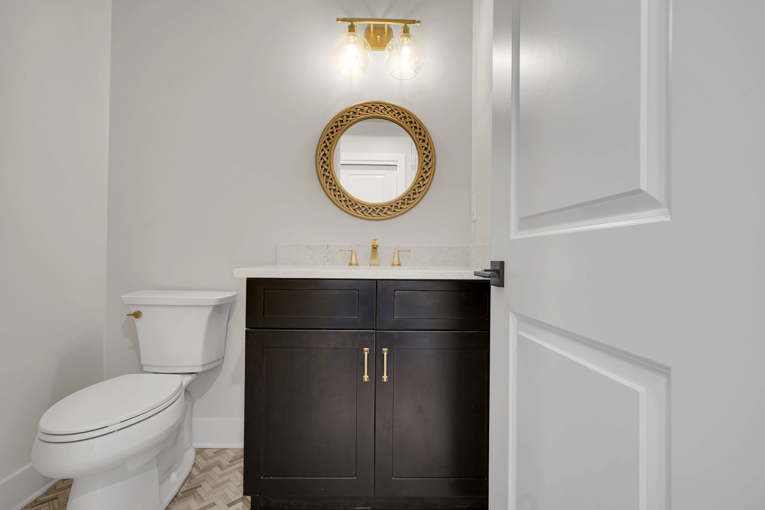 A small bathroom with a white toilet, dark vanity with gold handles, white countertop, round gold-framed mirror, and two-bulb gold light fixture above the sink. The floor has a herringbone pattern.