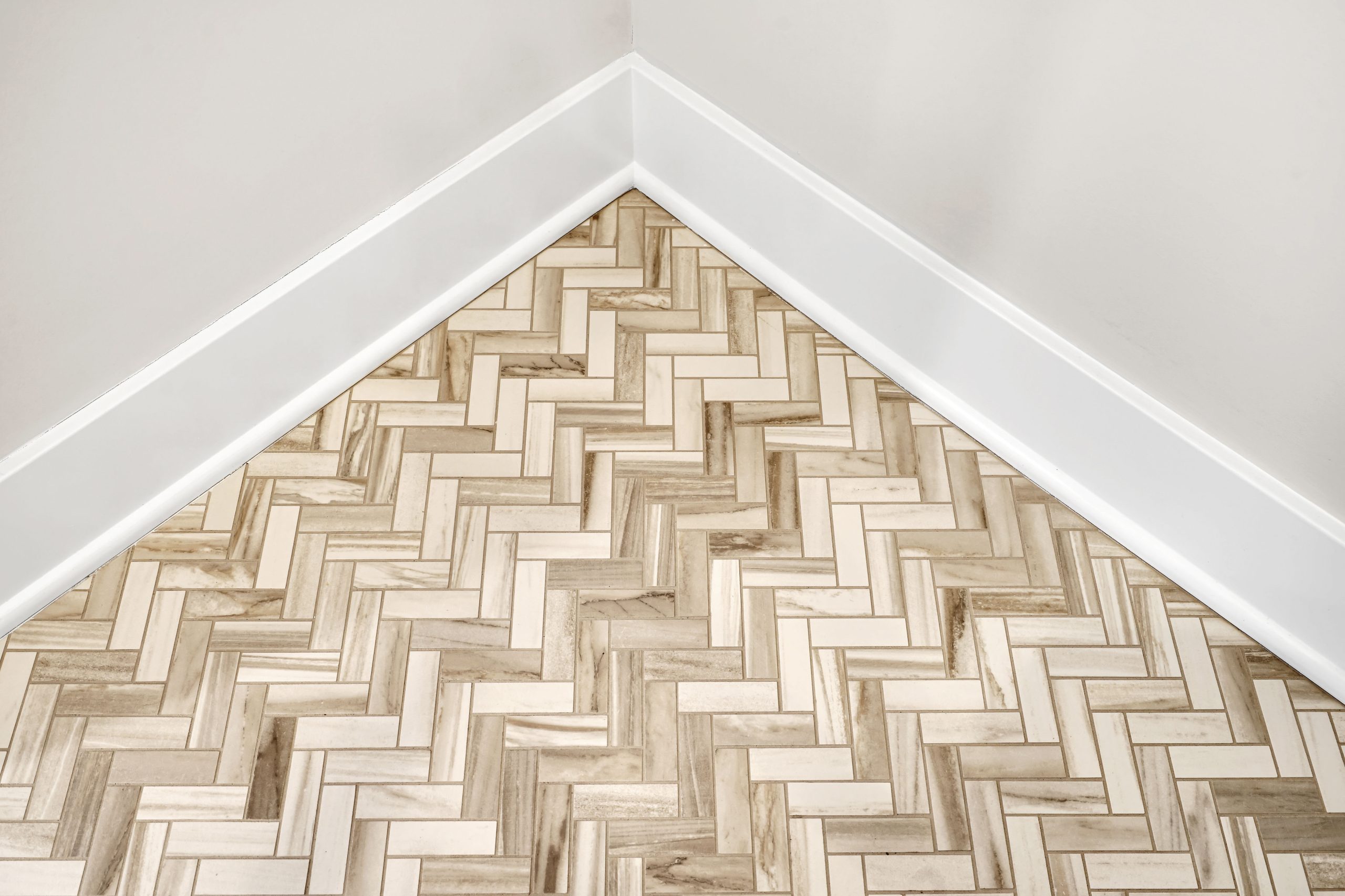 A corner of a room with light beige walls, white baseboards, and a light wood-patterned herringbone tile floor.