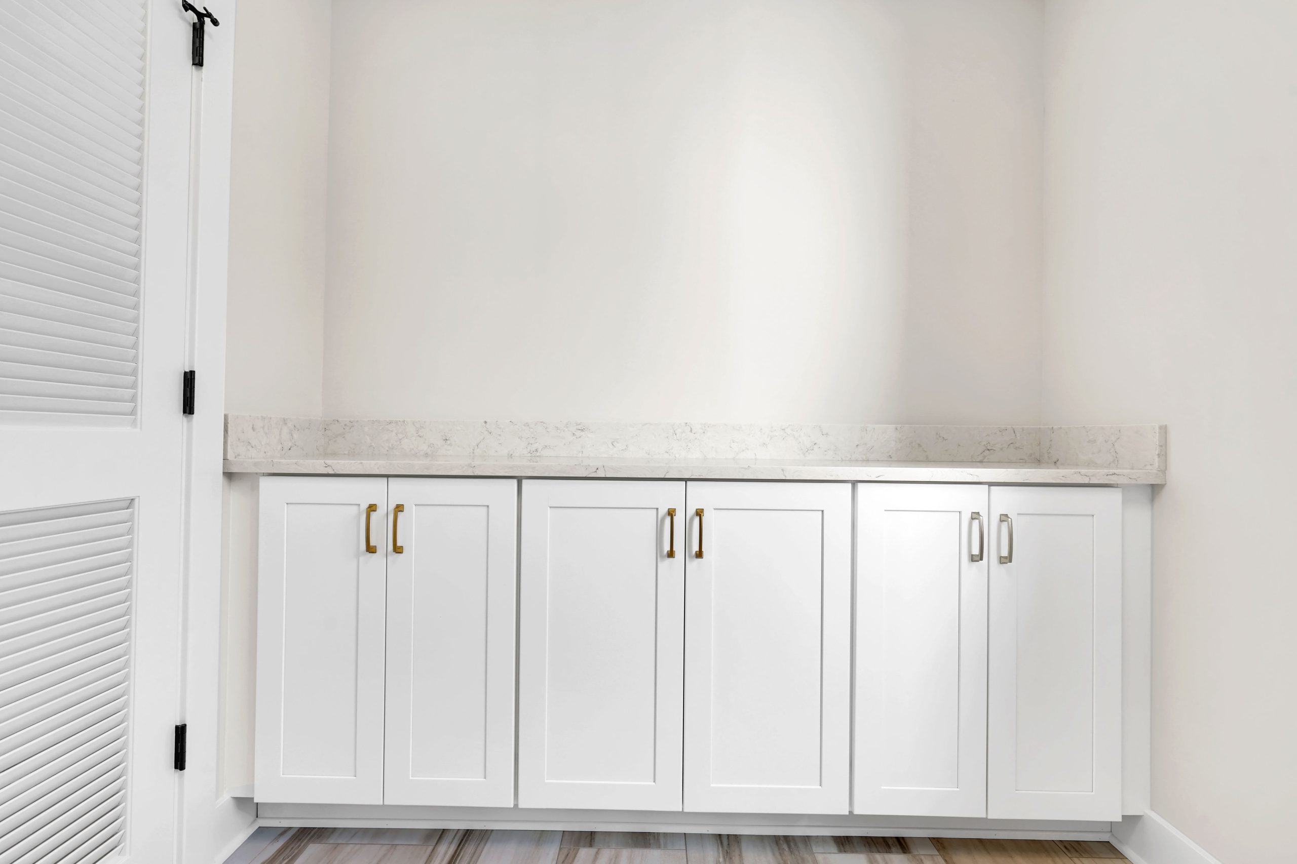 White cabinets with gold handles and a light marble countertop are set against a plain white wall. The floor is wood-patterned, and part of a white door with slats is visible on the left side of the image.