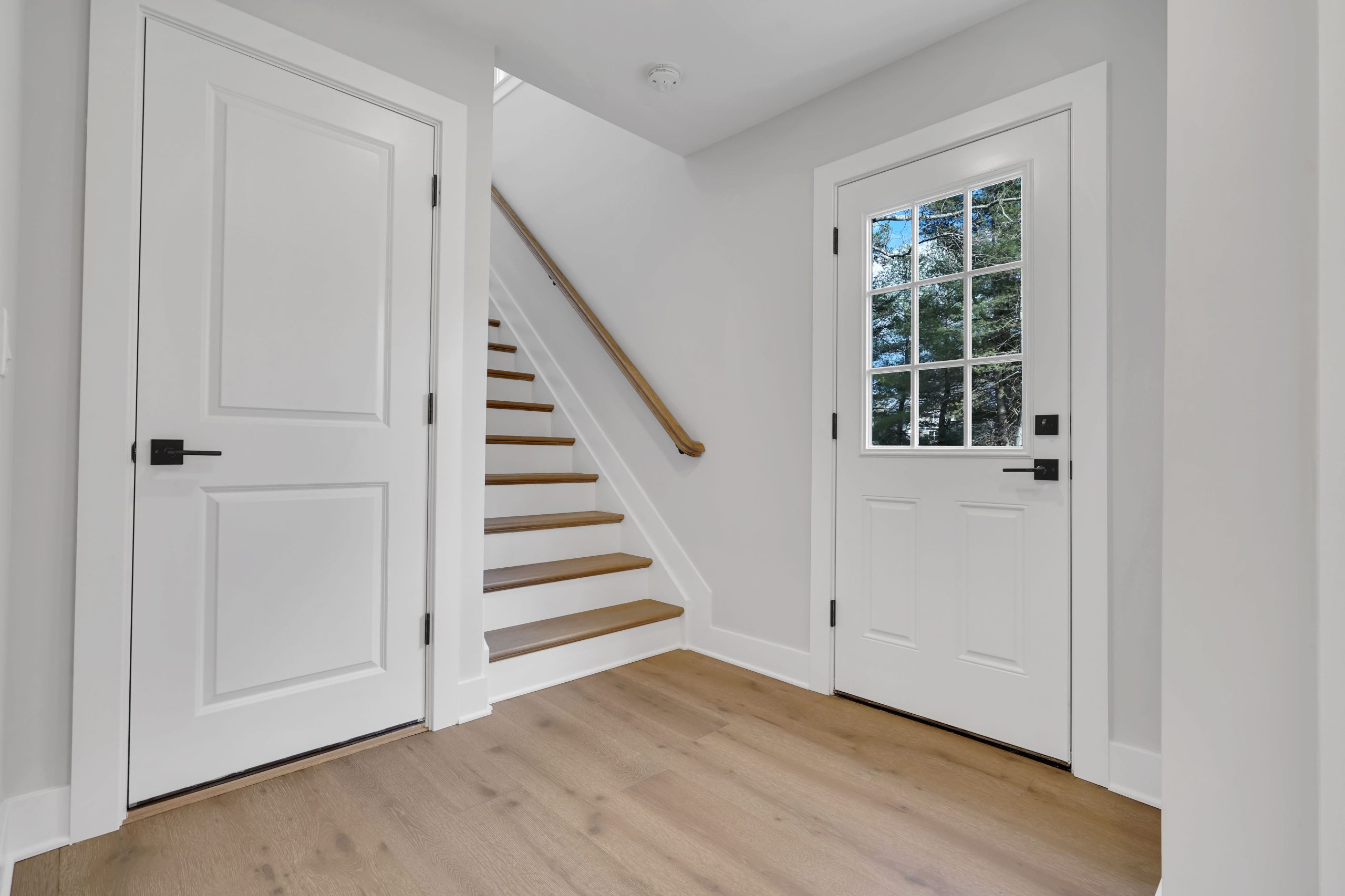A bright, modern entryway with light wood flooring, white walls and doors, a staircase with wooden steps and a simple wooden handrail, and a door with glass panes showing greenery outside.