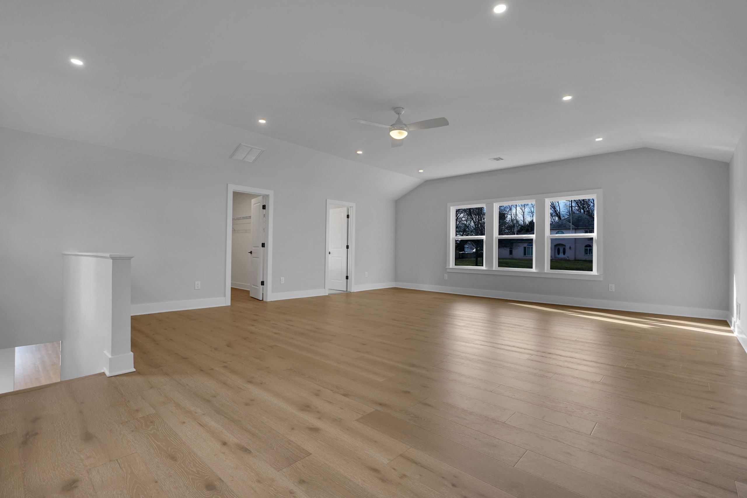 Spacious, empty room with light wood floors, white walls, recessed ceiling lights, a ceiling fan, and three large windows allowing natural light in. Two doors and a staircase are visible on the left side.