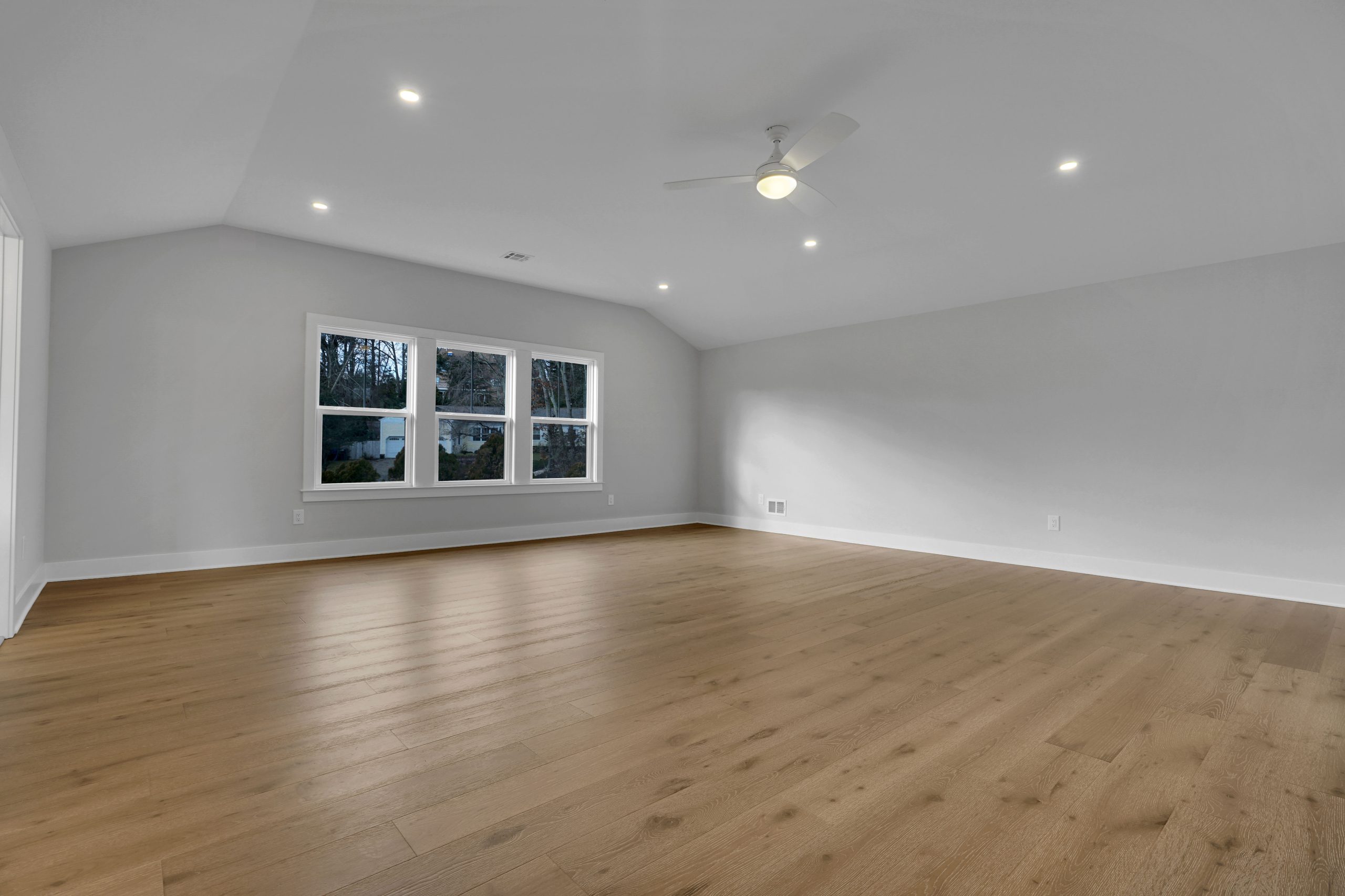 Spacious empty room with light wood flooring, white walls, large windows, recessed ceiling lights, and a ceiling fan. Natural light enters through the windows, highlighting the clean and modern design.