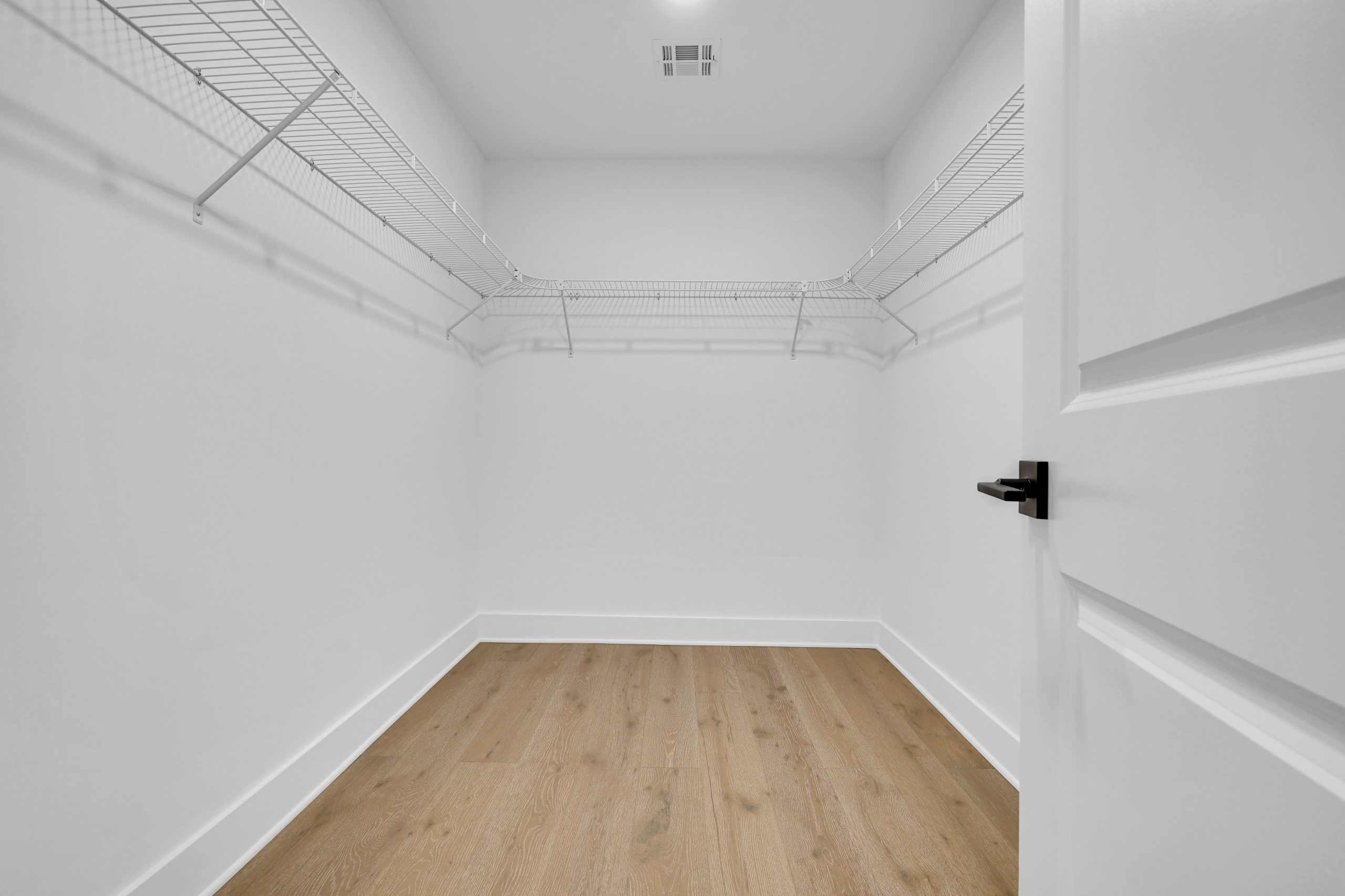 Empty walk-in closet with white walls, open door, wooden floor, and wire shelving along two walls. Ceiling air vent visible, and overall lighting is bright and clean.