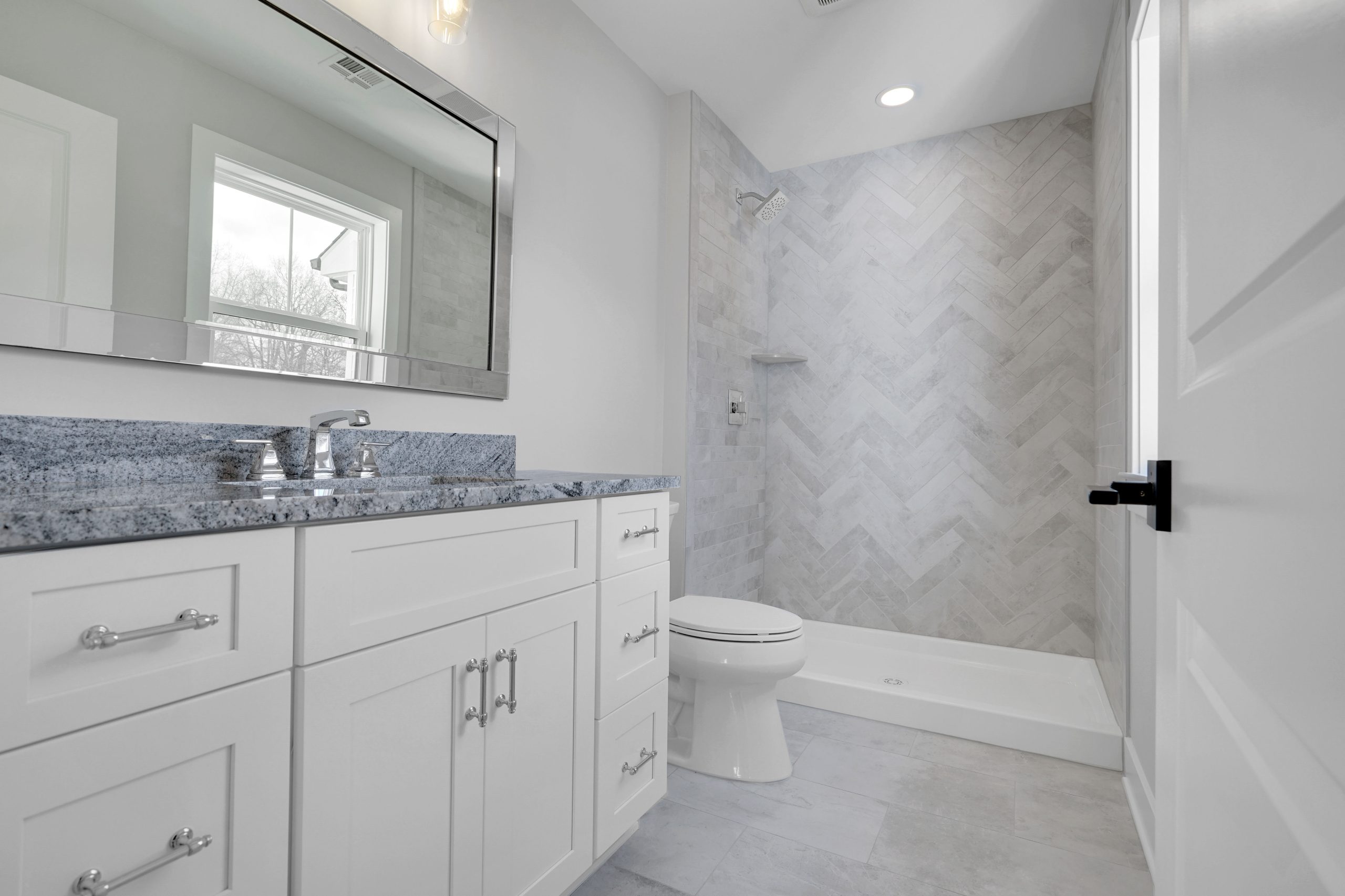 Modern bathroom with white cabinets, blue granite countertop, large mirror, silver fixtures, toilet, and a walk-in shower featuring light gray herringbone tile. Natural light enters from a window above the vanity.