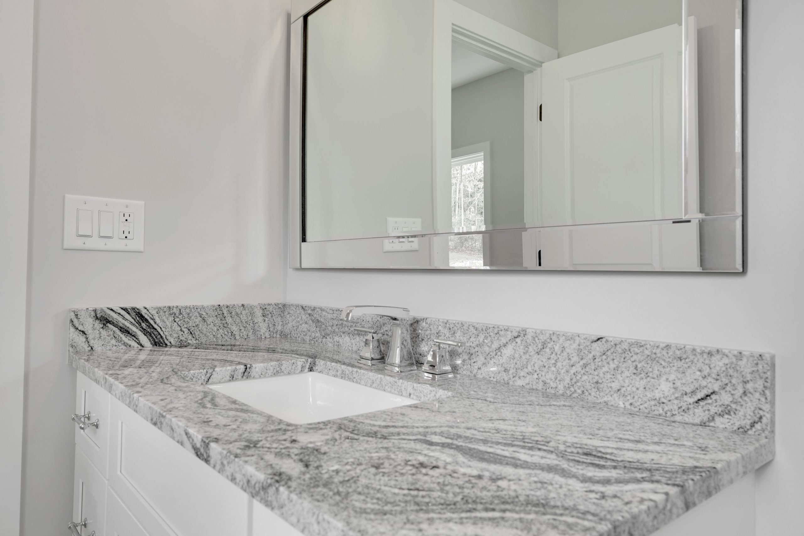 Modern bathroom vanity with a gray and white marble countertop, under-mount sink, silver faucet, white cabinetry, and a large rectangular mirror reflecting a doorway and window. Walls are light gray.
