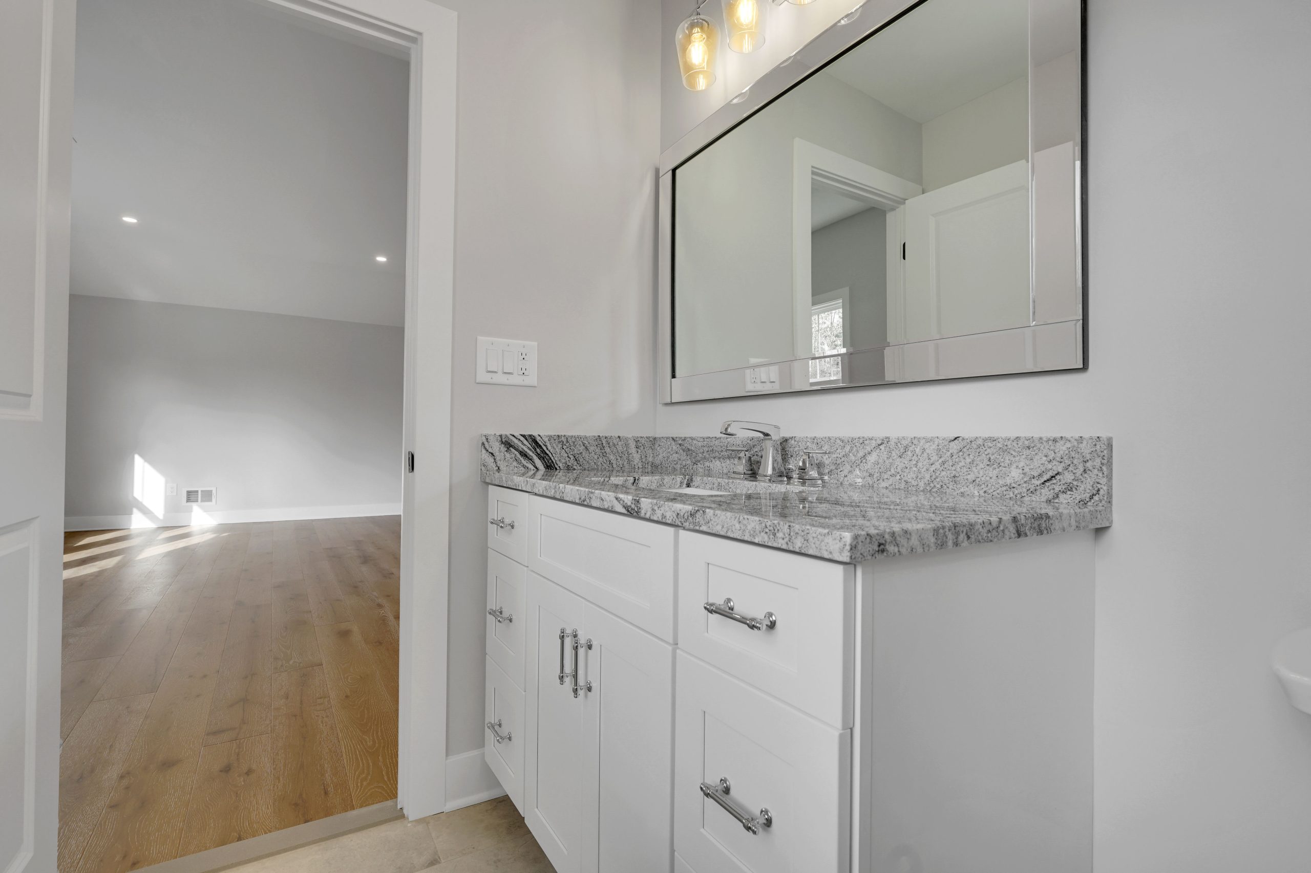 Modern bathroom with a white vanity, gray marble countertop, rectangular mirror, and three-bulb vanity light. An open door reveals an adjacent room with wood flooring and light gray walls.