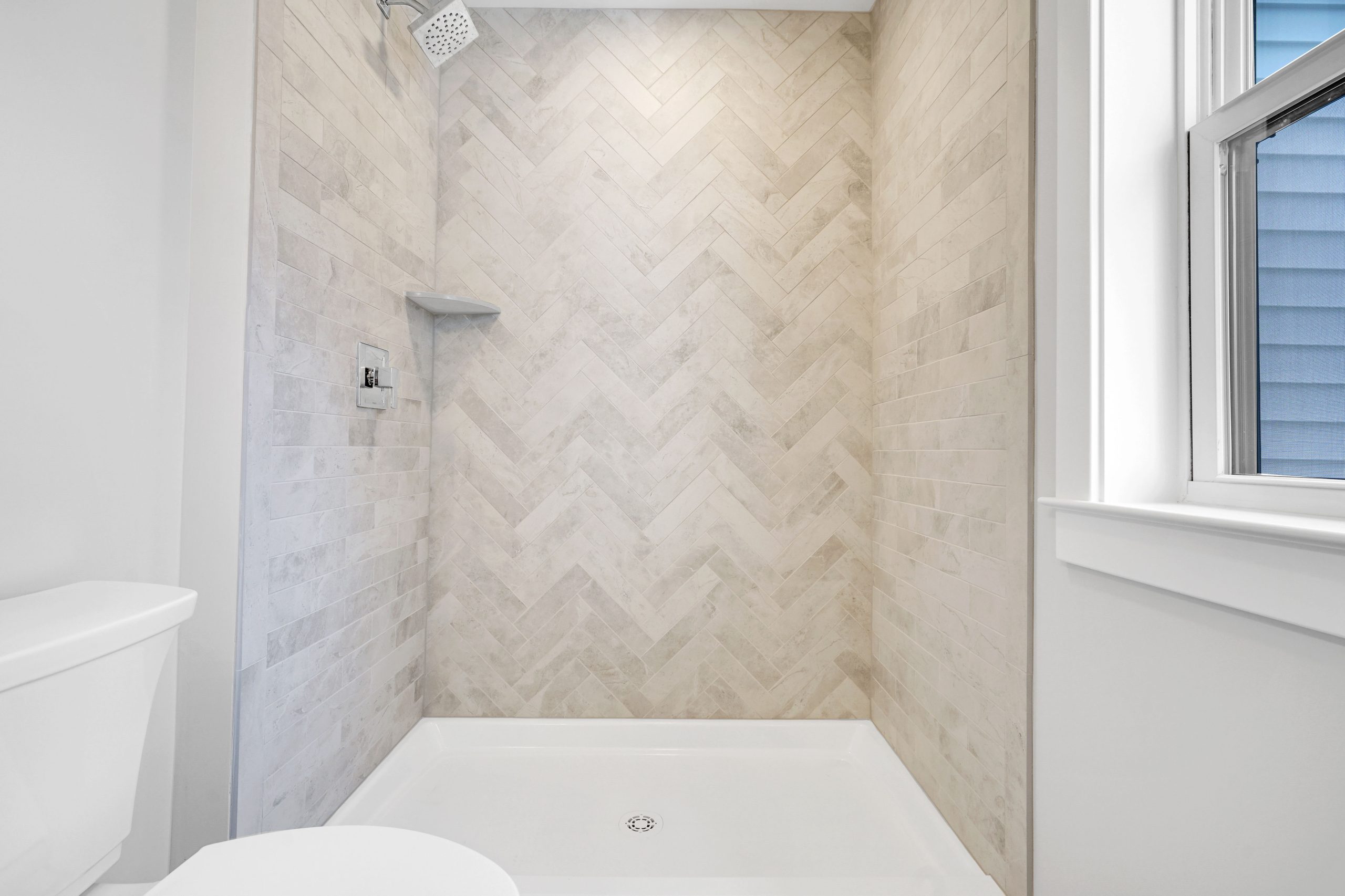 Modern shower with light beige herringbone tile pattern, built-in corner shelf, square showerhead, and a window on the right letting in natural light. White toilet partially visible on the left.