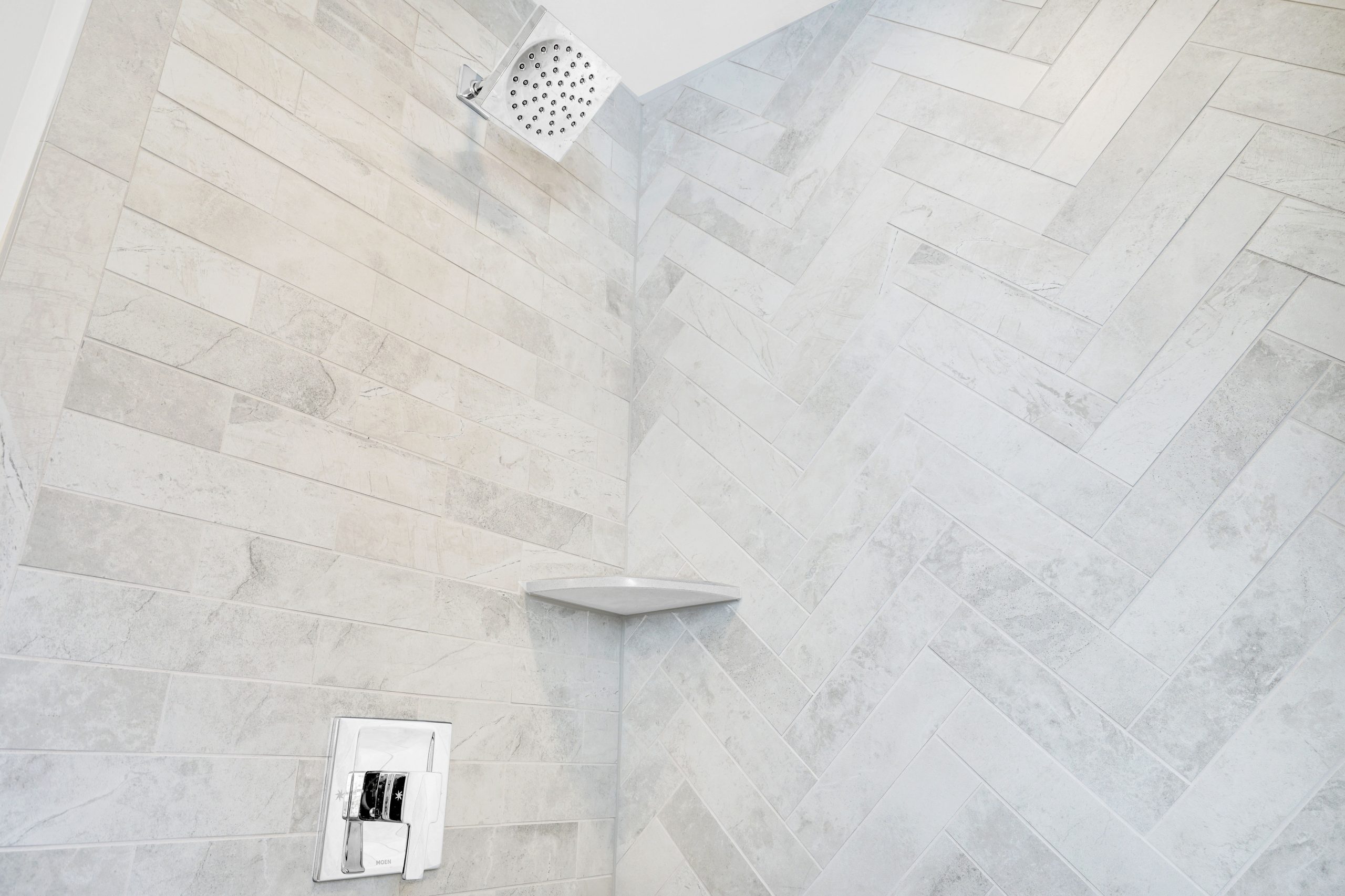 Modern shower with light gray herringbone-pattern tile walls, a square chrome showerhead at the top, a small corner shelf, and a sleek rectangular chrome control panel.
