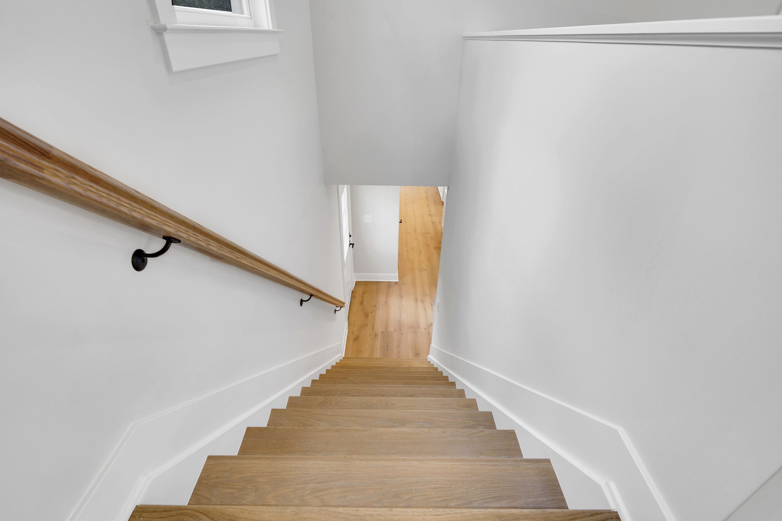 View looking down a straight, wooden staircase with a light brown handrail on the left, white walls on both sides, and light-colored wood flooring at the bottom. Natural light enters from a small window above.