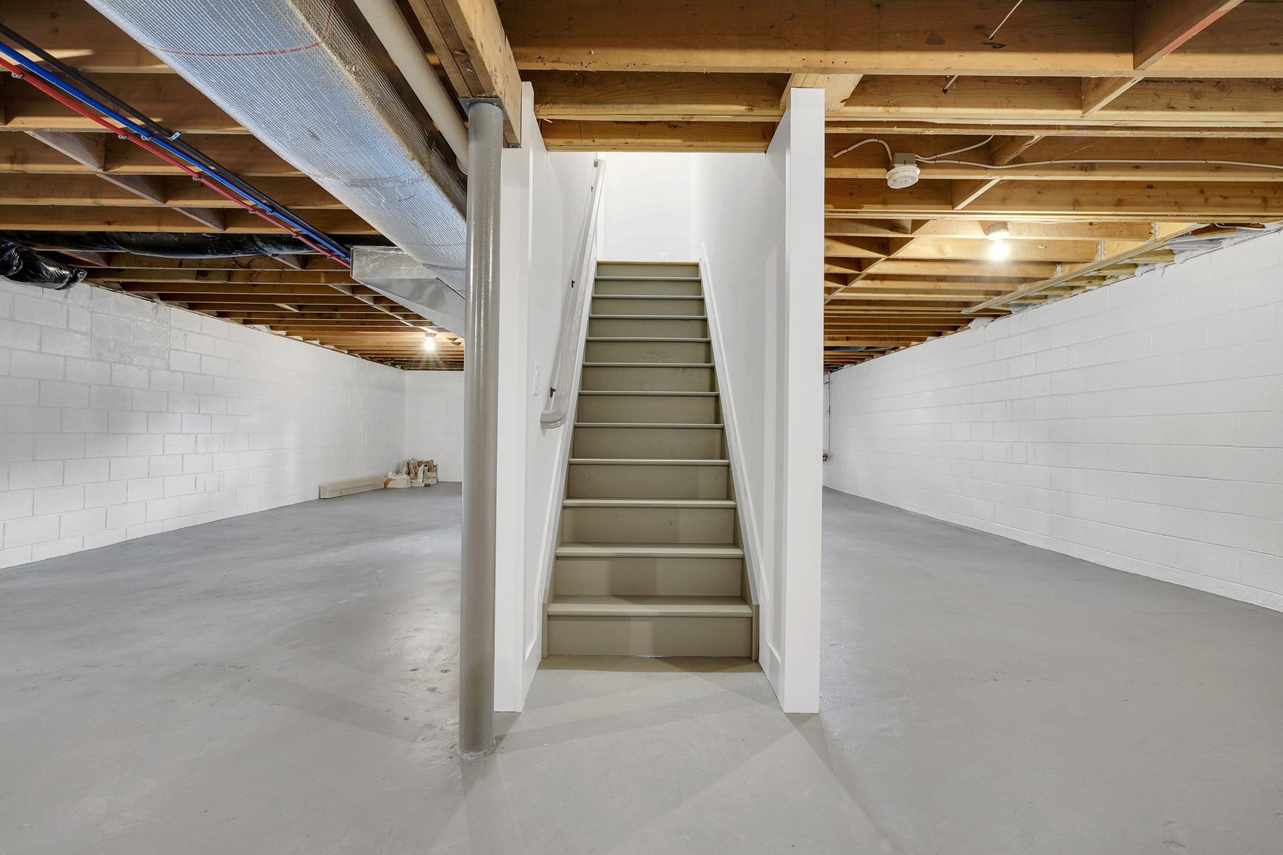 A clean, unfinished basement with exposed wooden ceiling beams, white painted cinder block walls, gray concrete floor, and a central staircase leading upstairs, dividing the basement into two spacious sections.