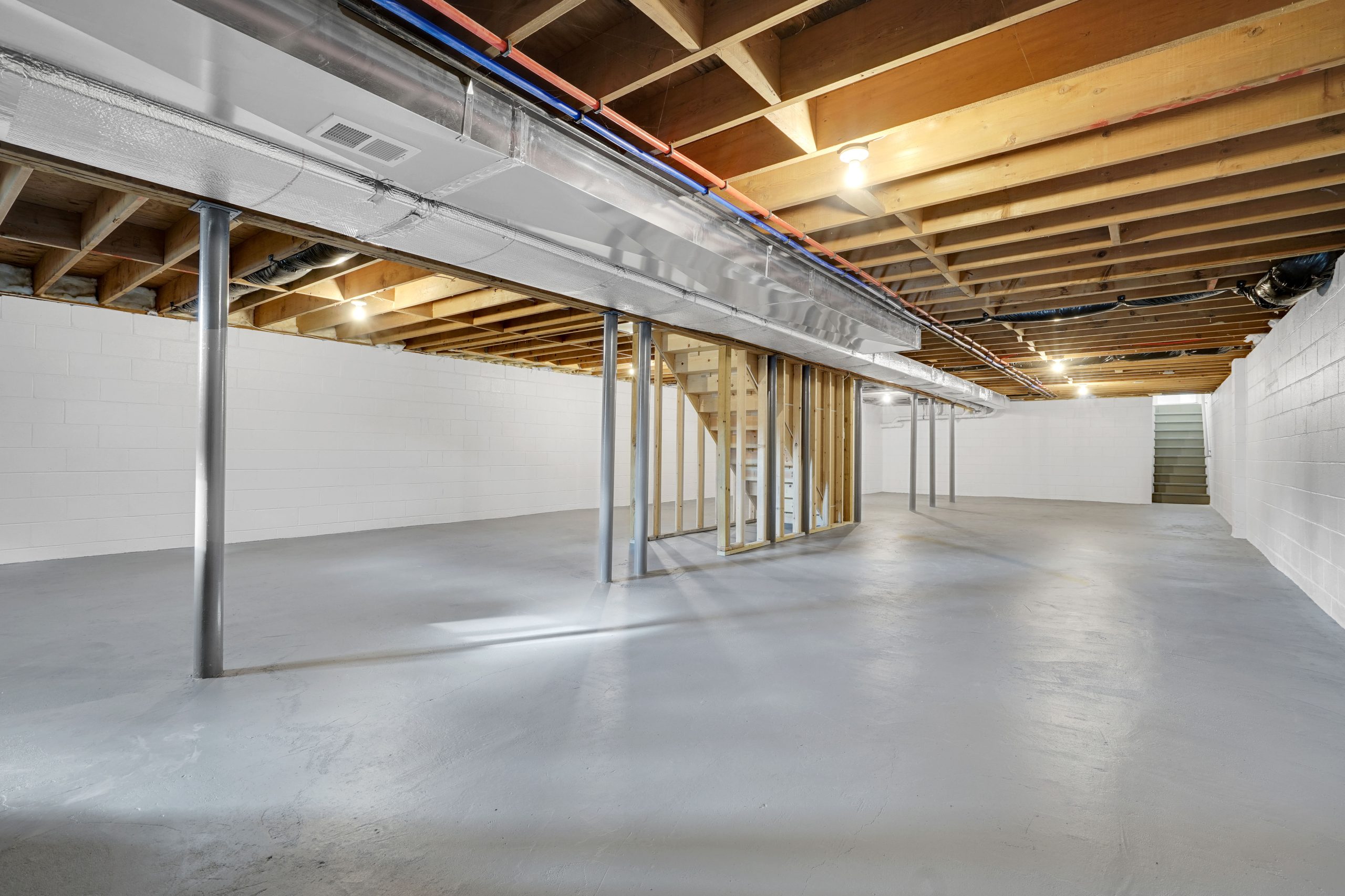 Unfinished basement with exposed wooden ceiling beams, white painted brick walls, grey concrete floor, metal support poles, visible ductwork, and a wooden staircase leading upstairs. The space is empty and brightly lit.