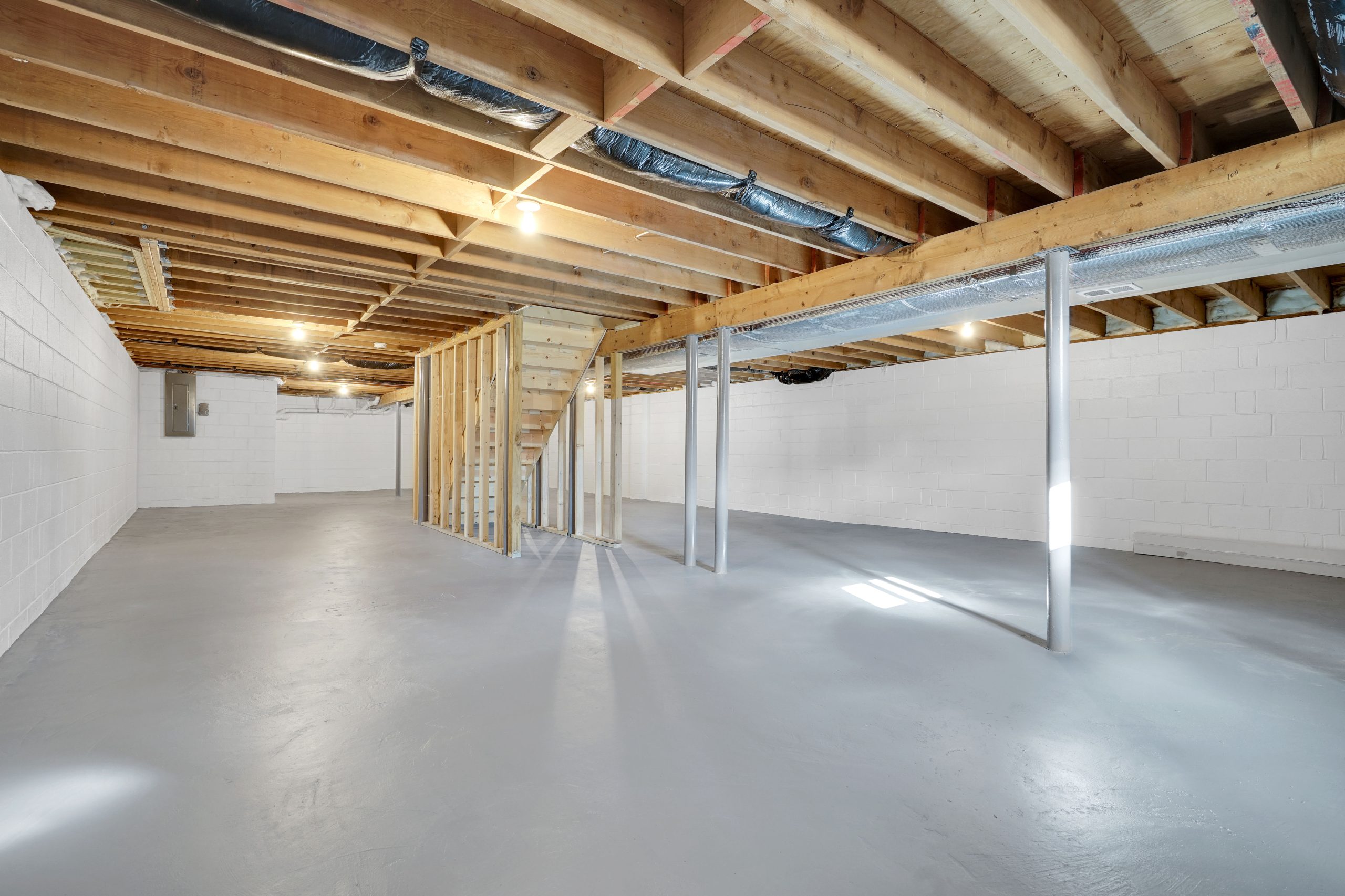 Unfinished basement with exposed wooden ceiling beams, support columns, concrete floor, white painted cinder block walls, and a wooden staircase leading upstairs. Natural light enters through small windows.