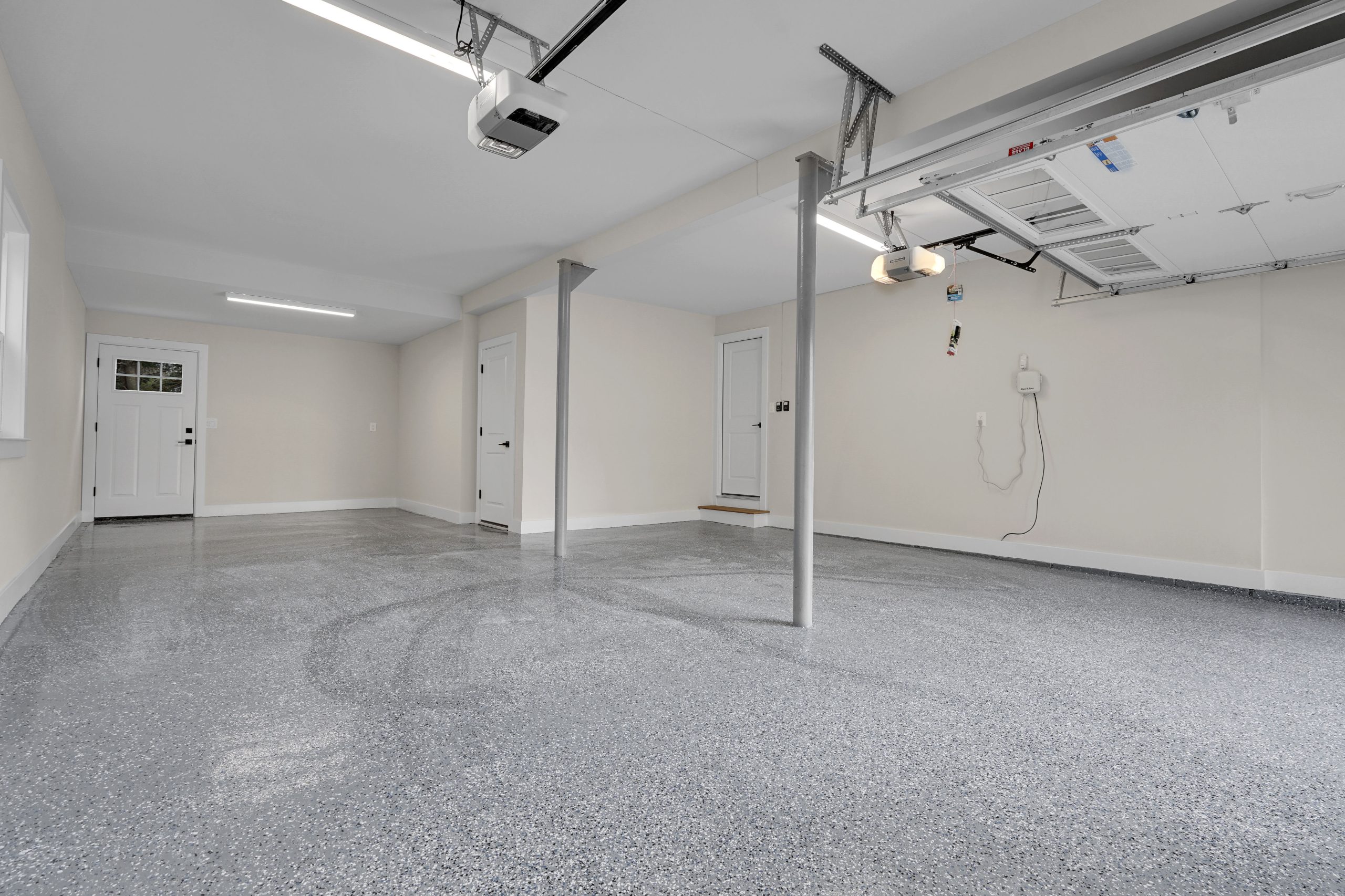 A clean, spacious garage with a speckled gray epoxy floor, white walls, overhead lighting, support beams, and open garage door. Several closed doors are visible, along with a wall-mounted control panel and windows.
