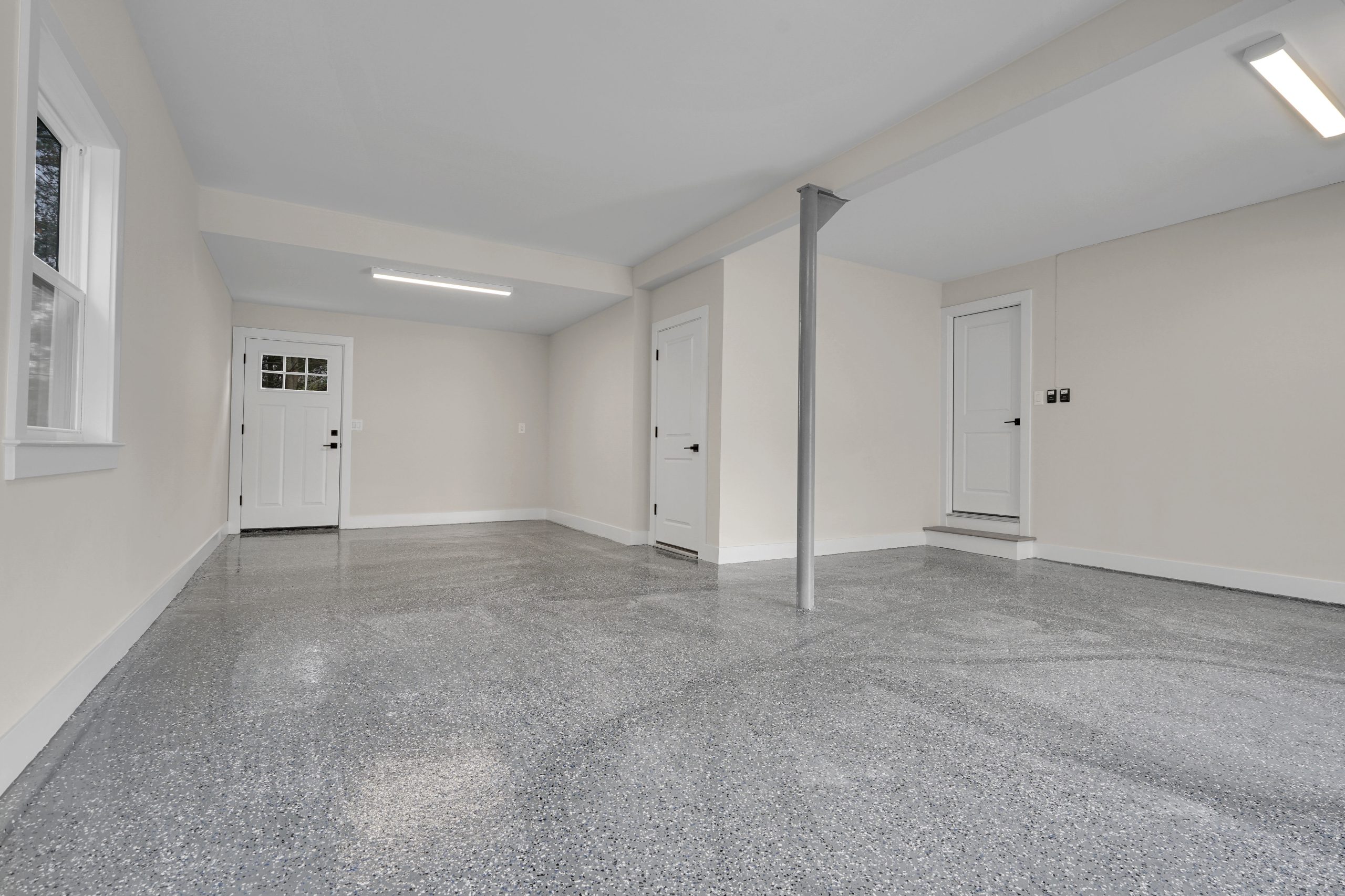 Bright, clean garage with light gray speckled epoxy floor, cream-colored walls, two white doors, a window on the left, and a metal support post near the center. Fluorescent ceiling lights provide illumination.