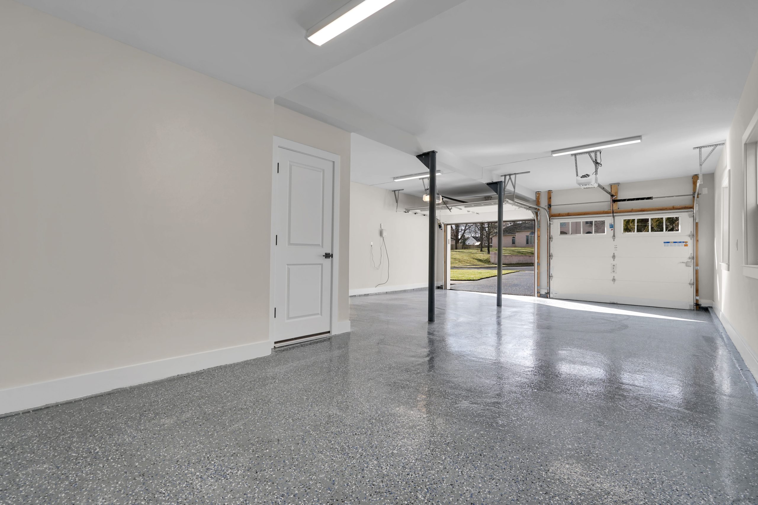 Clean, empty two-car garage with polished speckled gray floor, white walls, ceiling lights, a closed door on the left, and an open garage door showing a driveway and green lawn outside.