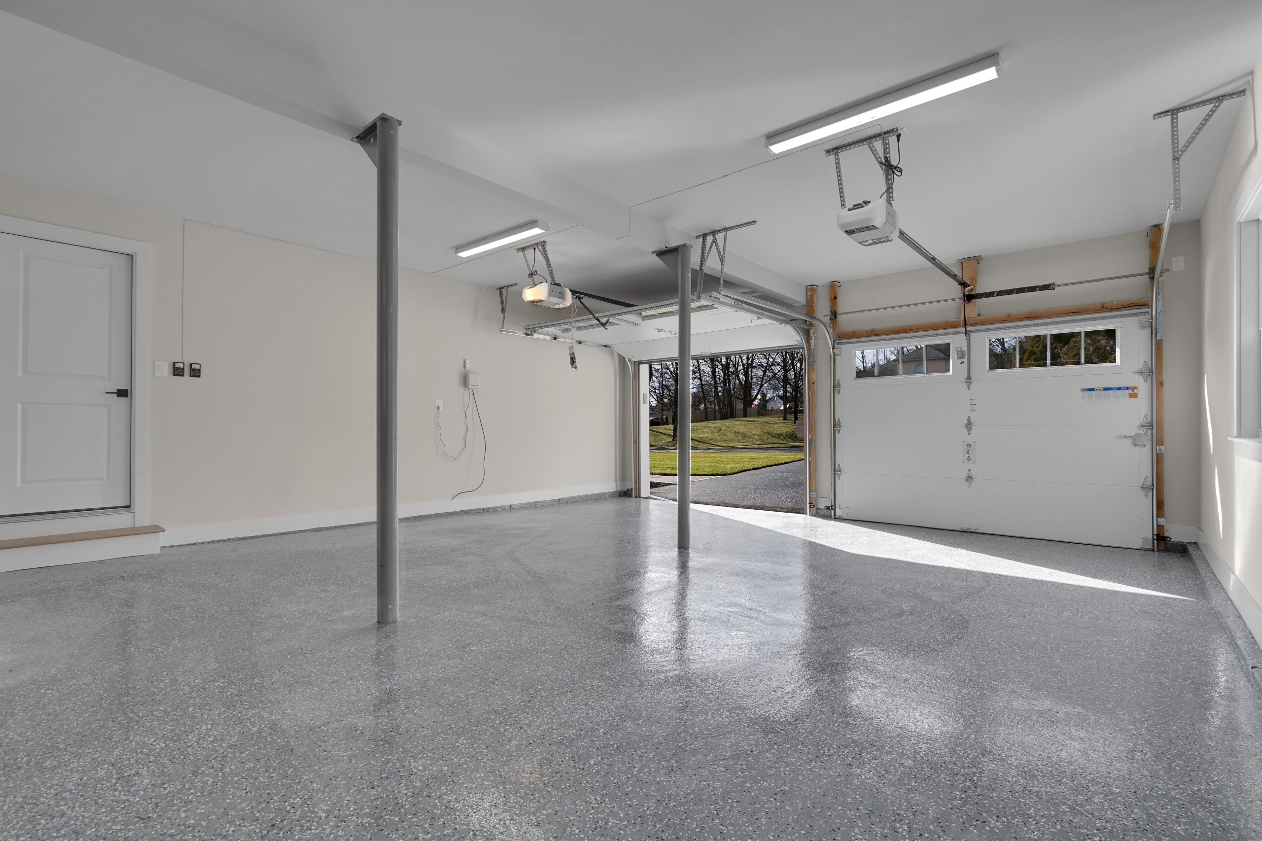 A clean, empty two-car garage with polished gray flooring, white walls, support columns, overhead lighting, and one garage door open to a driveway with trees visible outside.