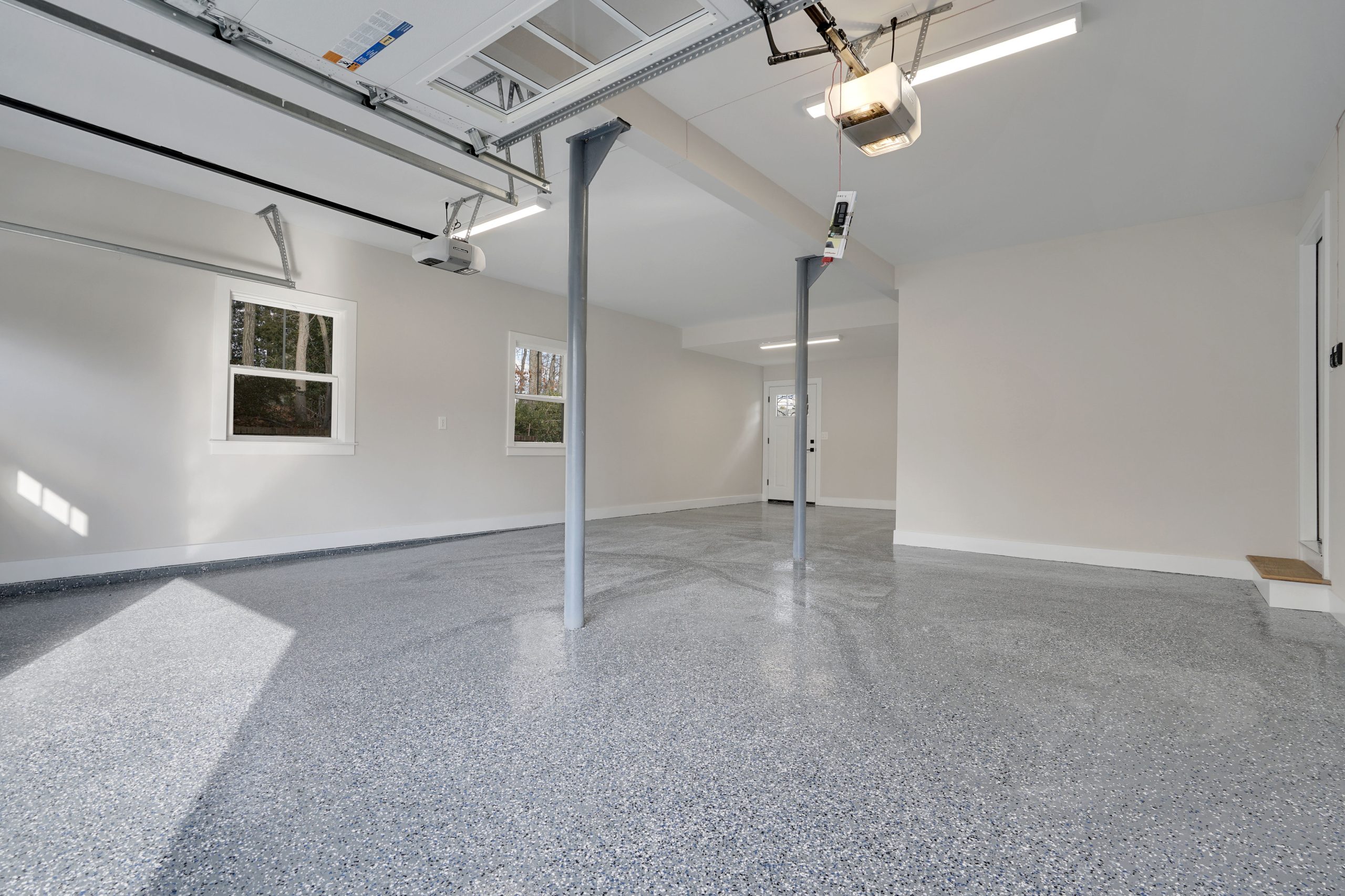 Clean, spacious two-car garage with speckled epoxy flooring, light-colored walls, two support columns, ceiling lights, two garage doors, windows, and a door at the back. Daylight shines through the windows.