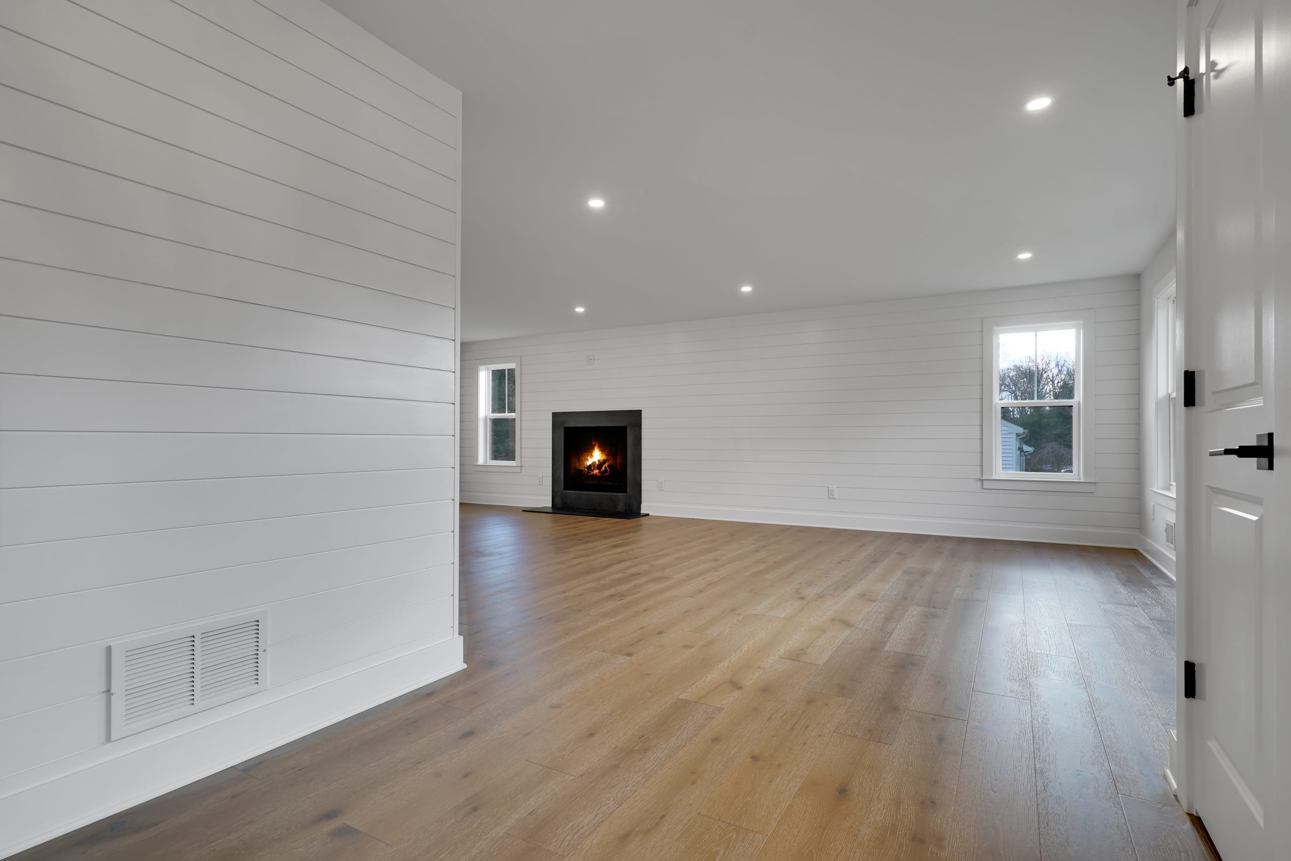 Spacious, modern living room with light wood flooring, white shiplap walls, recessed lighting, and a black fireplace. Two windows provide natural light. The room is empty and bright.