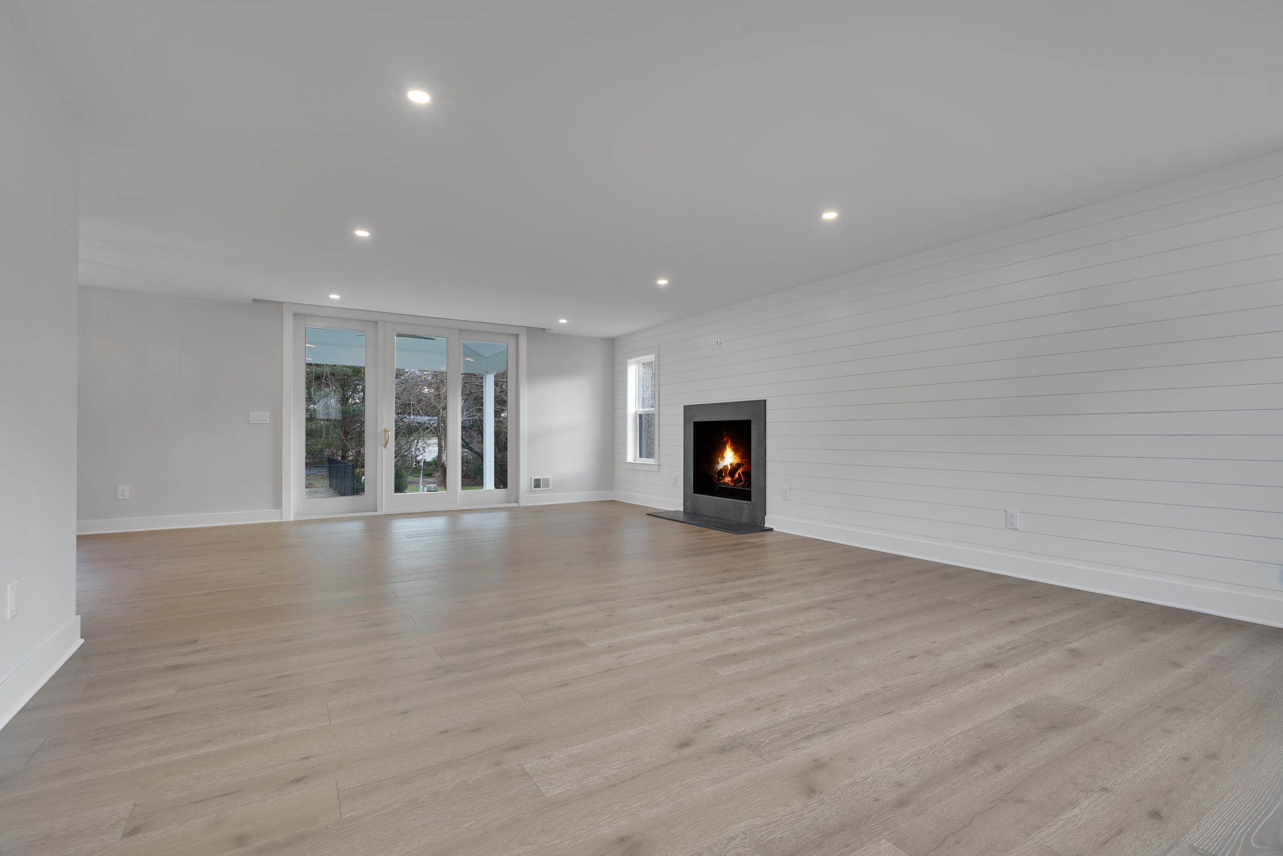 Spacious, empty living room with light wood floors, white shiplap walls, a modern black fireplace with a fire burning, recessed ceiling lights, and large glass doors leading to a backyard.