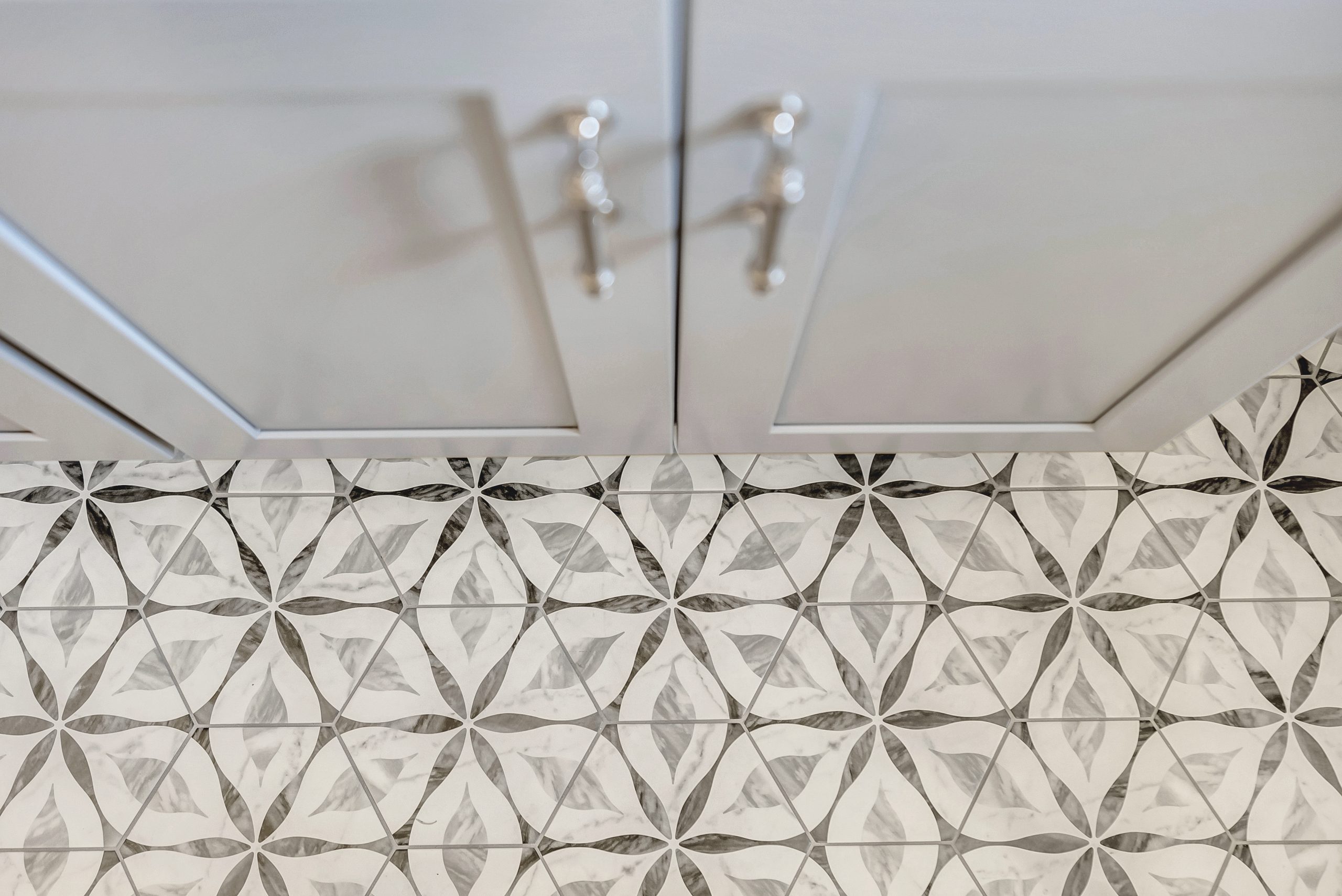 Gray cabinet doors with silver handles are seen from above, set against a floor with white tiles featuring a repeating geometric floral pattern in shades of gray and black.