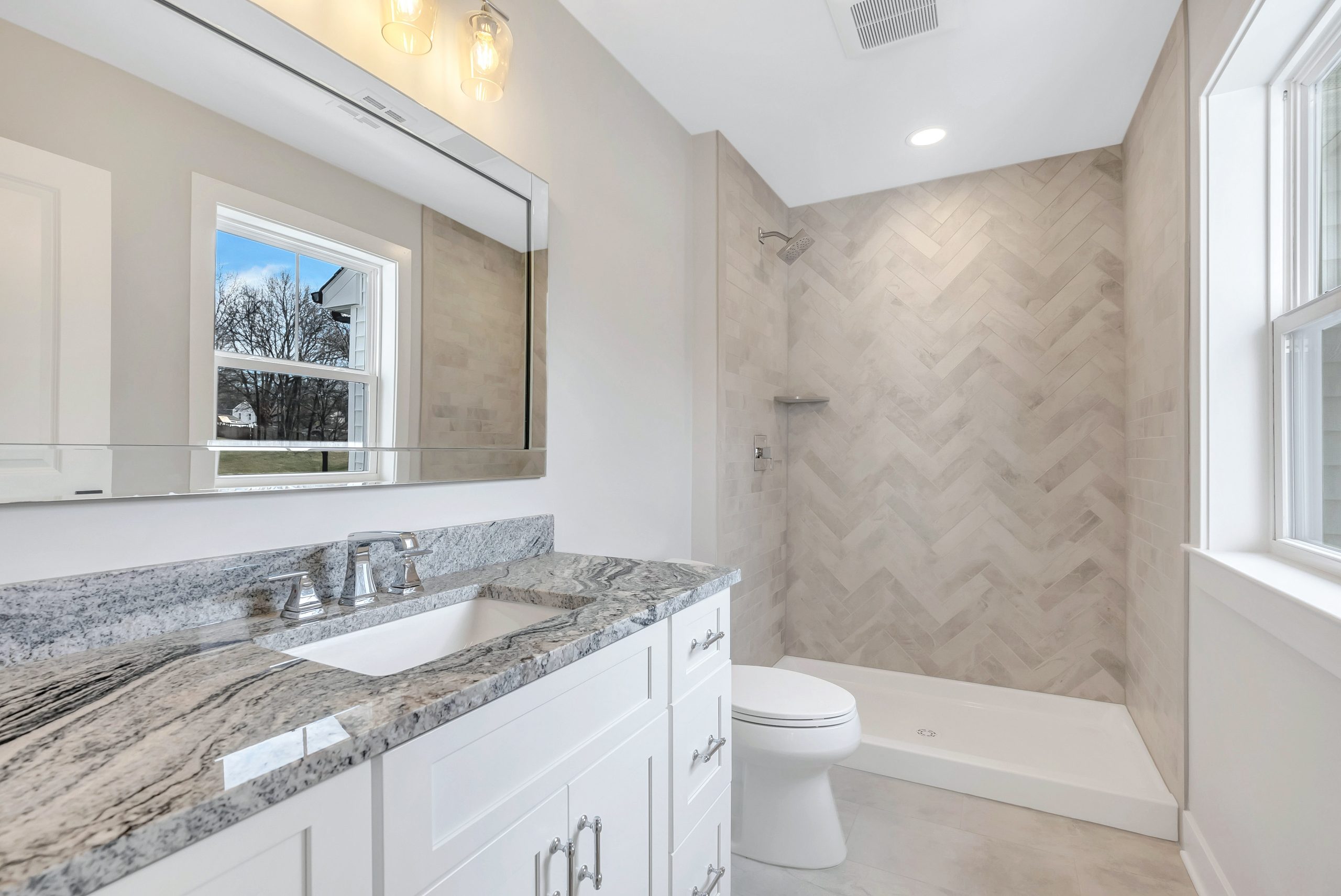 Modern bathroom with a white vanity, marble countertop, large mirror, and a walk-in shower featuring light herringbone tile. Natural light enters through two windows, creating a bright, clean atmosphere.