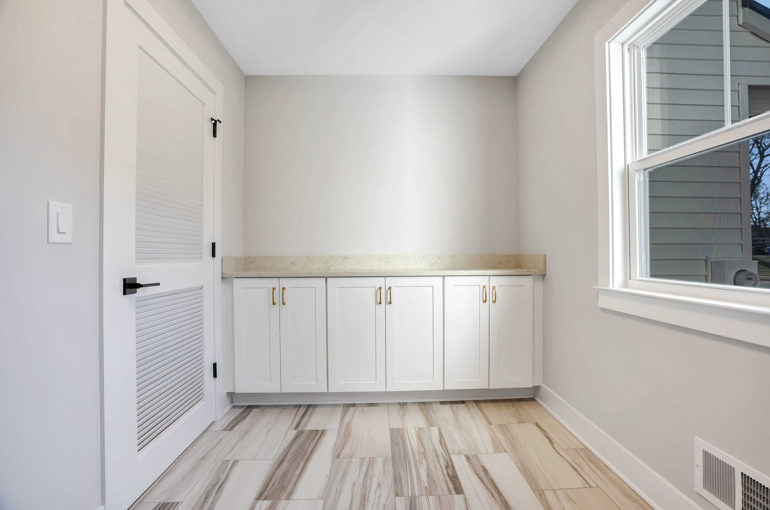A bright, empty room with white cabinets, gold handles, light beige countertop, tiled floor, white walls, a white door with black hardware, and a large window letting in natural light.